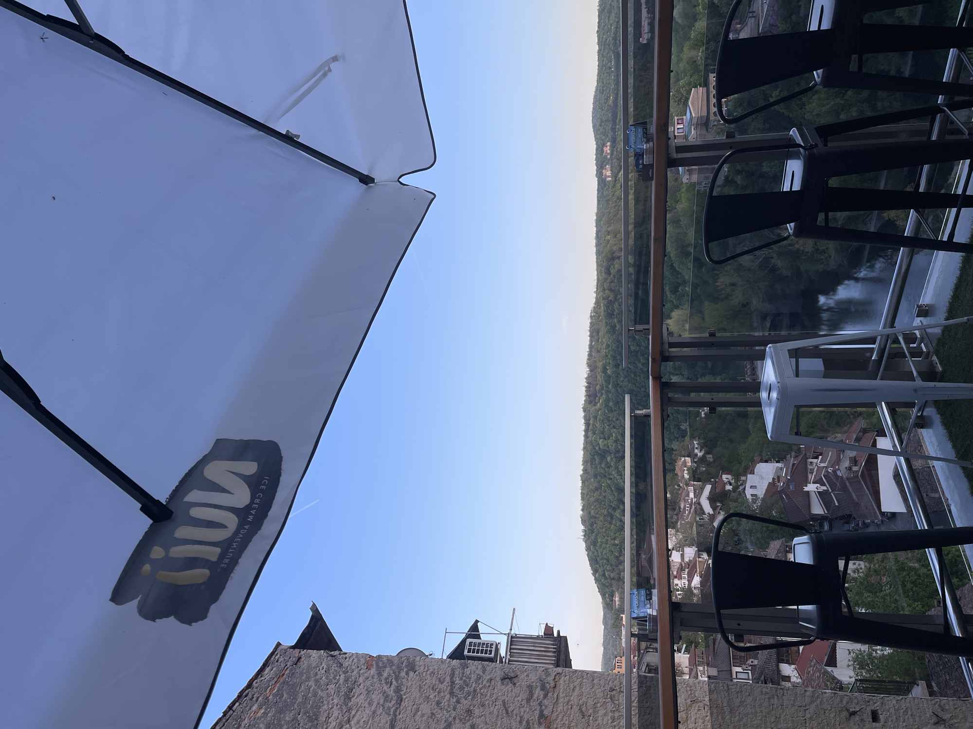 The image displays a panoramic view from an elevated outdoor terrace or balcony, overlooking a picturesque town and natural landscape. In the immediate foreground, a large white umbrella is partially visible on the left, with its underside showing a dark, stylized logo and the reversed text "ICE CREAM ADVENTURE". To the right, a series of modern bar stools, mostly black with one white, are arranged behind a clear glass and metal railing.
Beyond the railing, the scene opens to a European town, characterized by numerous buildings with distinctive red-tiled roofs nestled within a valley. A river, likely the Yantra River, flows through the heart of the landscape, bordered by dense, tree-covered hills that stretch to the horizon. The sky is clear and bright blue, suggesting fair weather and possibly late afternoon or early evening. The setting is consistent with a scenic viewpoint in Veliko Tarnovo, Bulgaria. No people are visible, emphasizing the serene and expansive vista.