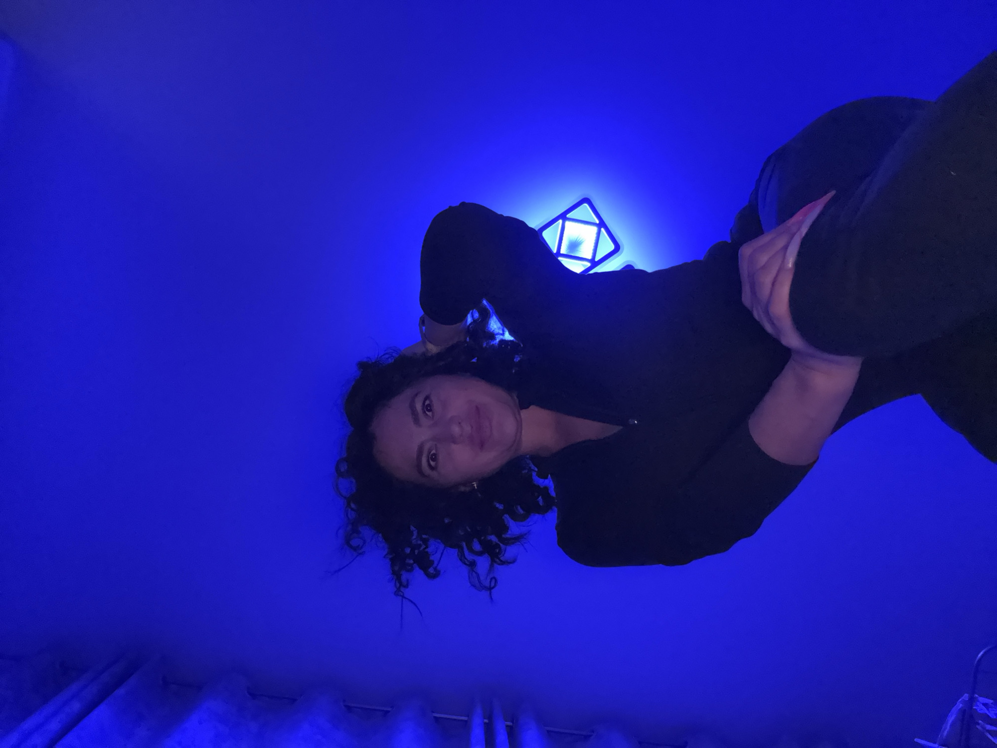 A woman with curly dark hair, wearing a black long-sleeved shirt and dark pants, is positioned centrally in the frame. The image appears to be taken from a low angle, looking up at the woman. Her face is slightly angled toward the viewer. Above her is a square-shaped light fixture emitting a bright white light. The background is primarily a solid blue color, possibly indicating a wall or ceiling with blue lighting. At the bottom edge of the frame, there are hints of what appears to be a curtain rod. The scene's location cannot be determined as Kauchuk Stantsiya, Russia, as there are no identifiable markers.