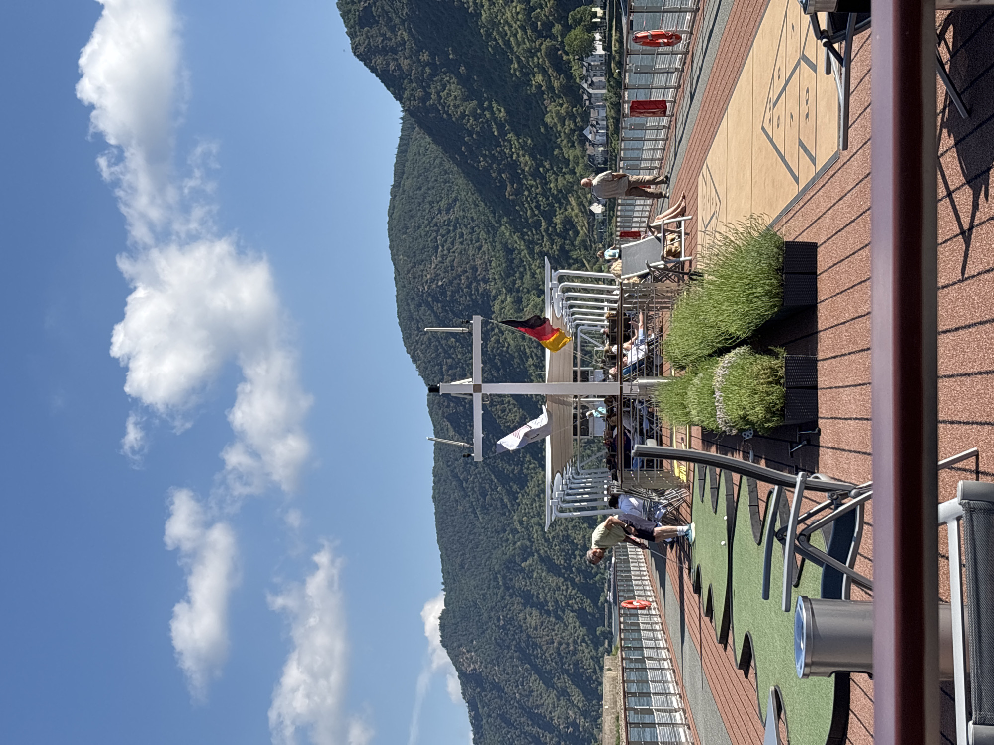 The image depicts a sunny day on a cruise ship deck in Kestert, Germany. The foreground shows a miniature golf putting area with green turf and metal putters. A person in a light green shirt and dark shorts is visible on the putting green. Further back, a shuffleboard court is marked on the deck. Several people are seated in lounge chairs, some under umbrellas, enjoying the scenery. A German flag is prominently displayed on a flagpole.
In the background, a steep, tree-covered mountainside rises dramatically, with houses visible nestled along its base. The Rhine River is implied to be nearby, as suggested by the cruise ship setting and the riverside town. The sky is bright blue with fluffy white clouds, indicating clear and pleasant weather. The sun is casting distinct shadows from the railings and furniture, suggesting it is midday. The overall atmosphere appears relaxed and leisurely, with passengers enjoying the scenic views of the German landscape.