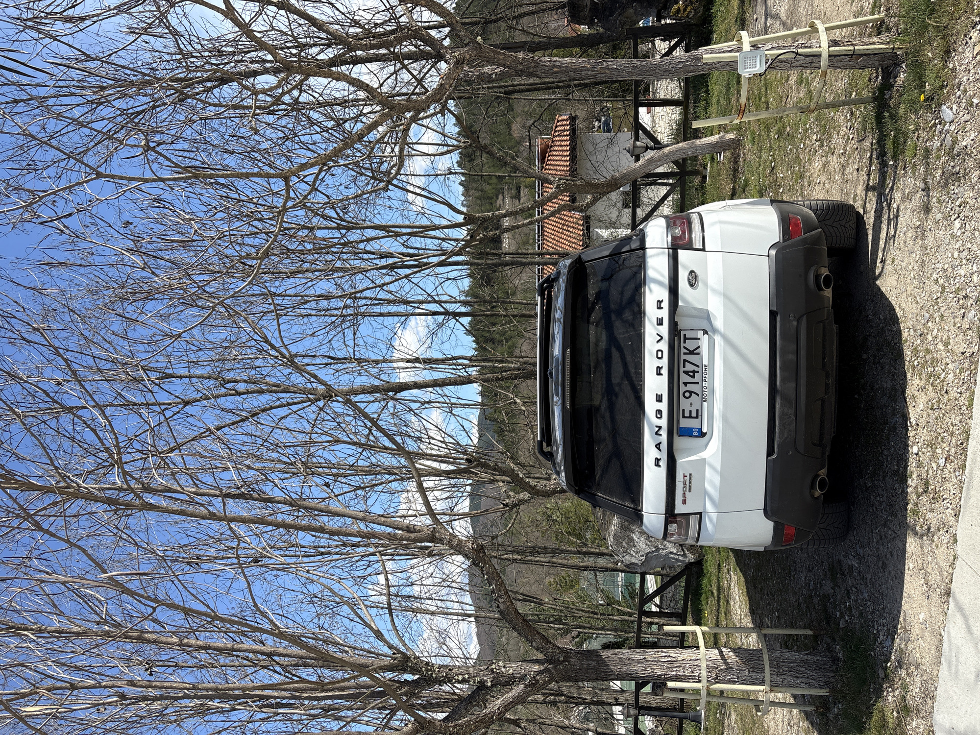 The image shows the rear of a white Range Rover Sport parked outdoors. The license plate reads "E 9147 KT" and indicates it is from Bulgaria. The car is parked on a gravel surface beside a concrete walkway and is partially obscured by bare tree branches. In the background, a glimpse of a building with a red-tiled roof and mountainous terrain suggests a location within a hilly or mountainous region. The image appears to have been taken in Koprivlen, Bulgaria, based on the license plate information. The overall environment suggests a rural or semi-rural setting.