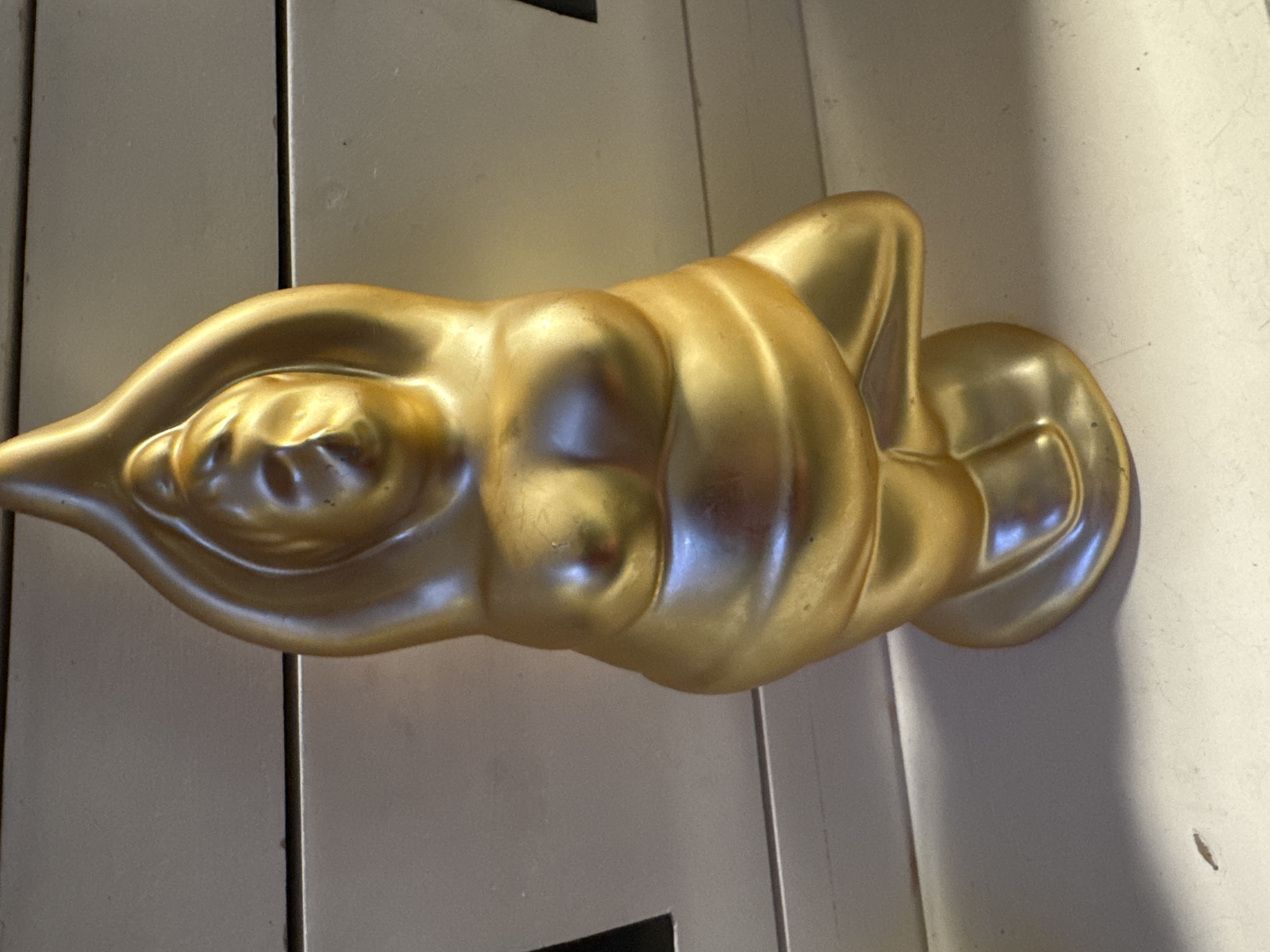 A gold-colored statue of a person in a yoga pose sits on a white surface. The statue is detailed, showing curves of the body and has a glossy finish. The background appears to be a light-colored wall with horizontal lines, possibly drawers or a textured surface. The lighting casts shadows and highlights on the statue. The image does not contain any elements or context that would indicate a location in Hallum, Netherlands.