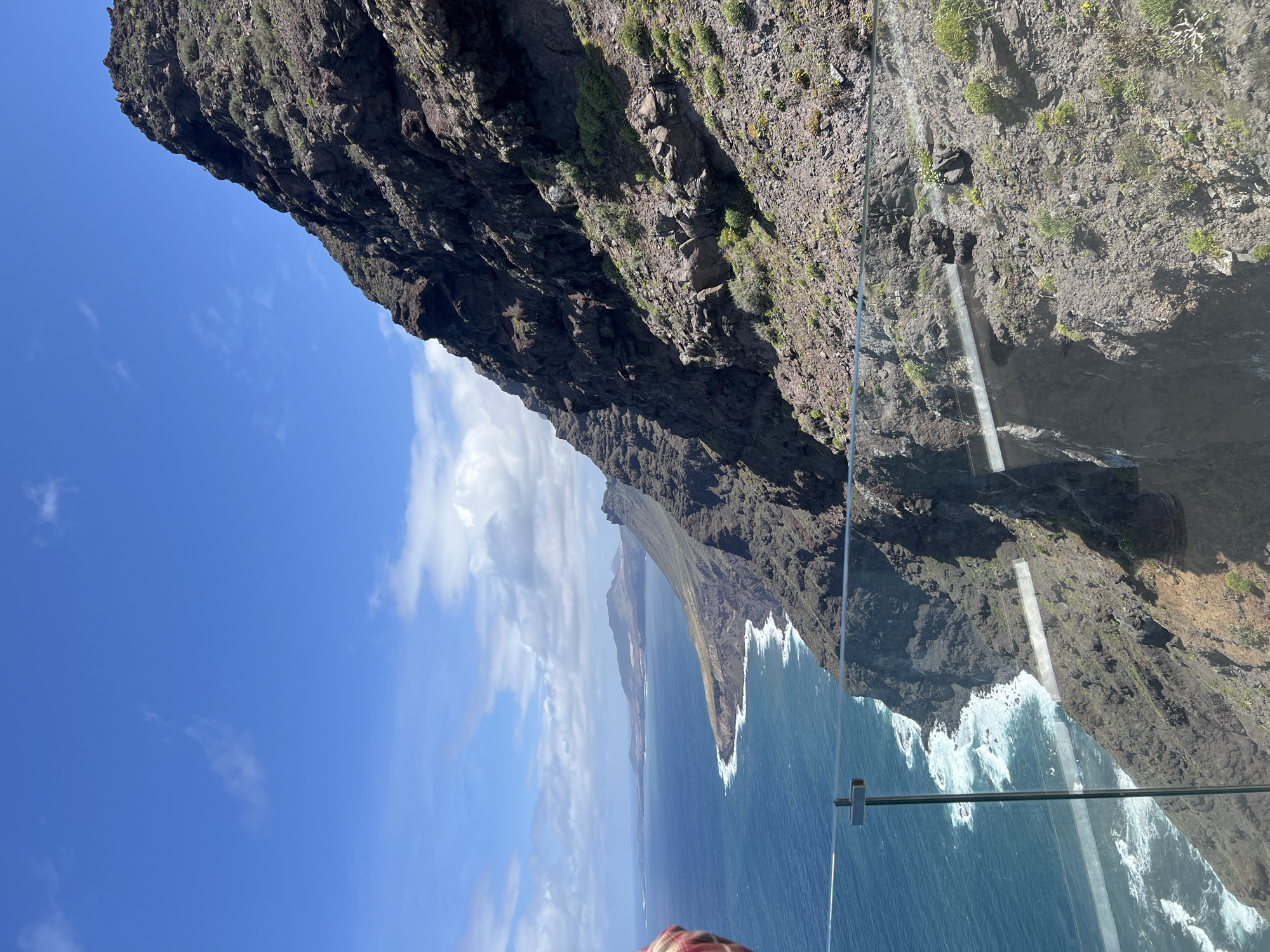 A daytime shot of a cliff in Tirma, Spain, is taken from a glass-enclosed lookout point. The sky is mostly blue with scattered clouds. The cliff is rugged and textured, with some sparse vegetation. The ocean below is a deep blue with white waves crashing against the base of the cliff. The glass enclosure is visible as a vertical barrier with metal support structures.