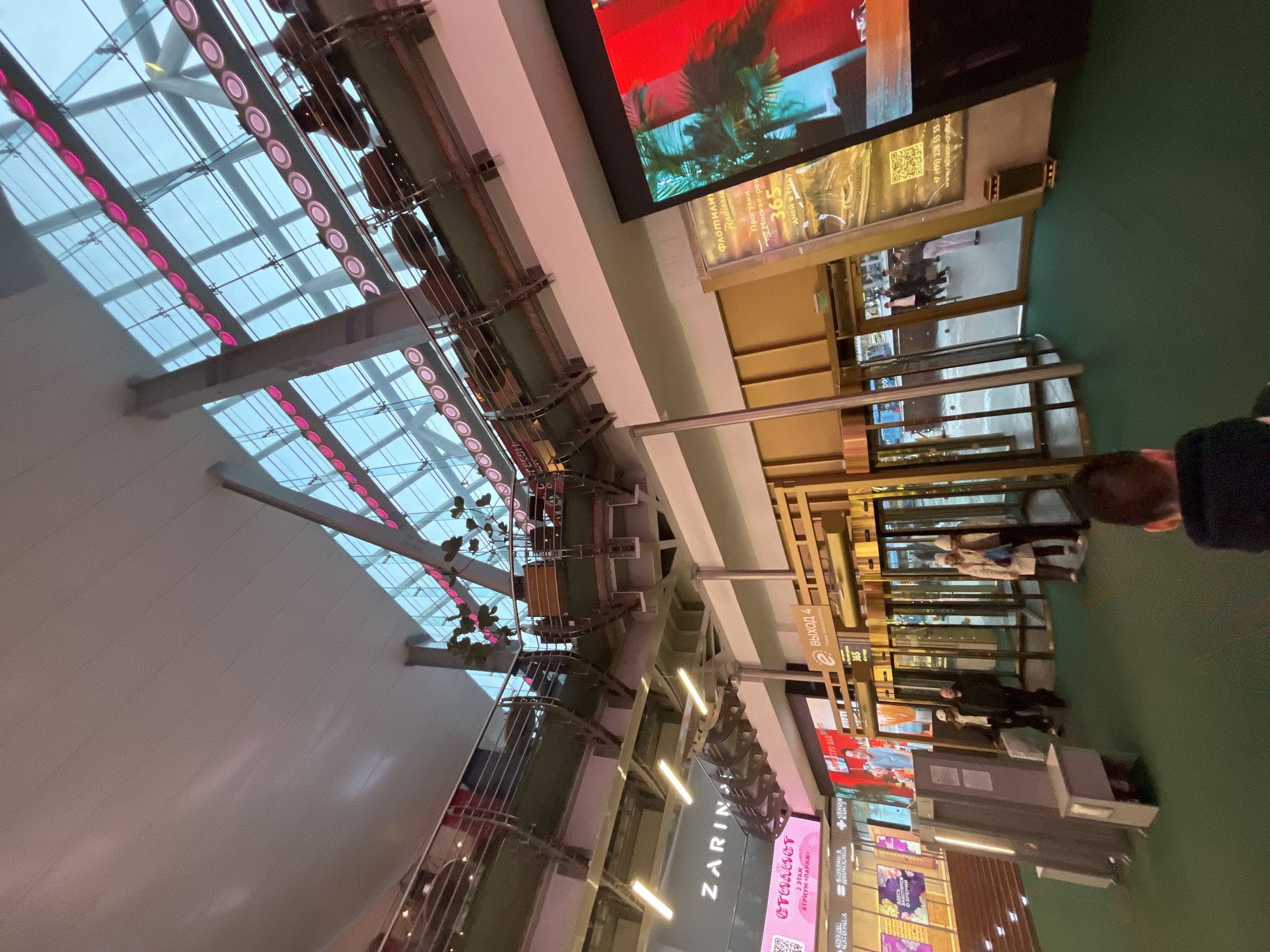 Here is a detailed description of the image:
**Location:** The image appears to be taken inside a shopping mall in Moscow, Russia. This is indicated by the presence of signage in Russian, including the names of brands like "Zarina" and "Radisson," and phrases like "Moscow-reka".
**Environment:** The image is taken looking upward from the mall's interior. The mall features a high, elaborate glass ceiling with pink lighting. There is a complex network of metal support beams for the ceiling.
**Subjects and Objects:** The mall has multiple levels, and we can see storefronts and advertising displays. There is a large LED screen displaying images, as well as multiple advertising signs. We can see the "Выход 4" (Exit 4) sign. There are mall entrances and people moving through the automatic doors.
**People:** The image includes a few people. The back of one person's head is visible in the foreground. Other people can be seen walking through the mall entrance.
**Actions:** The primary action is people walking through the mall and moving around the retail spaces. The environment suggests shopping and leisure activities are taking place.