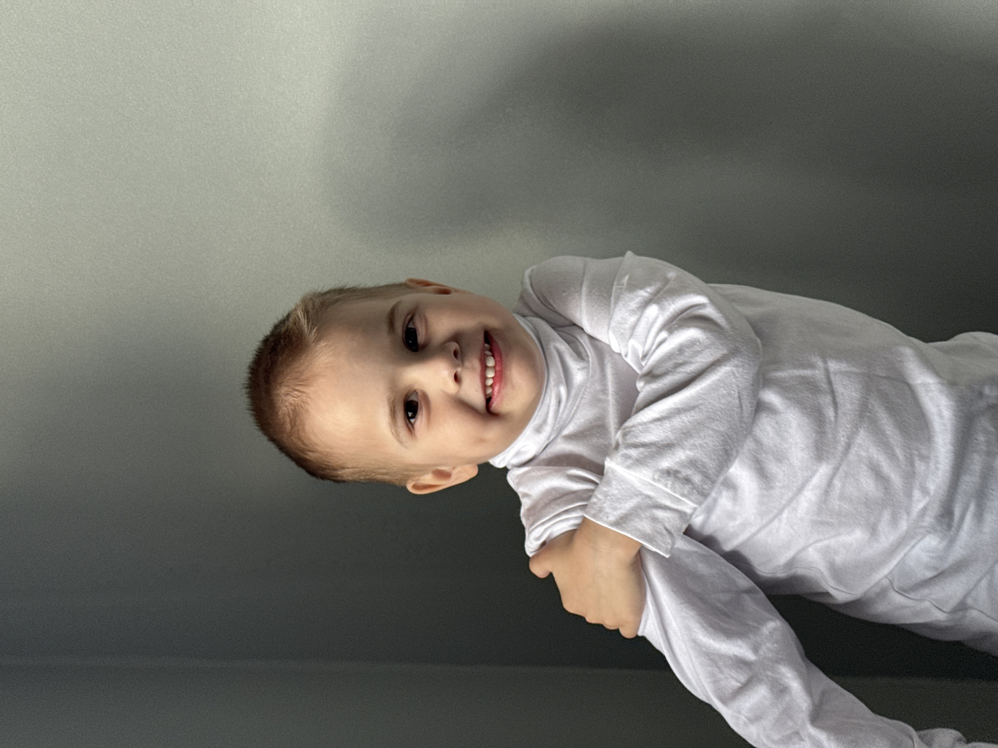 A young boy with short, light-brown hair smiles at the camera. He is wearing a white turtleneck long-sleeved shirt. His arms are crossed at his chest. The background is a gradient of gray, appearing to be a wall with light coming from above. The location is not discernible from the image and cannot be reliably determined to be in Chaikovsky, Russia.