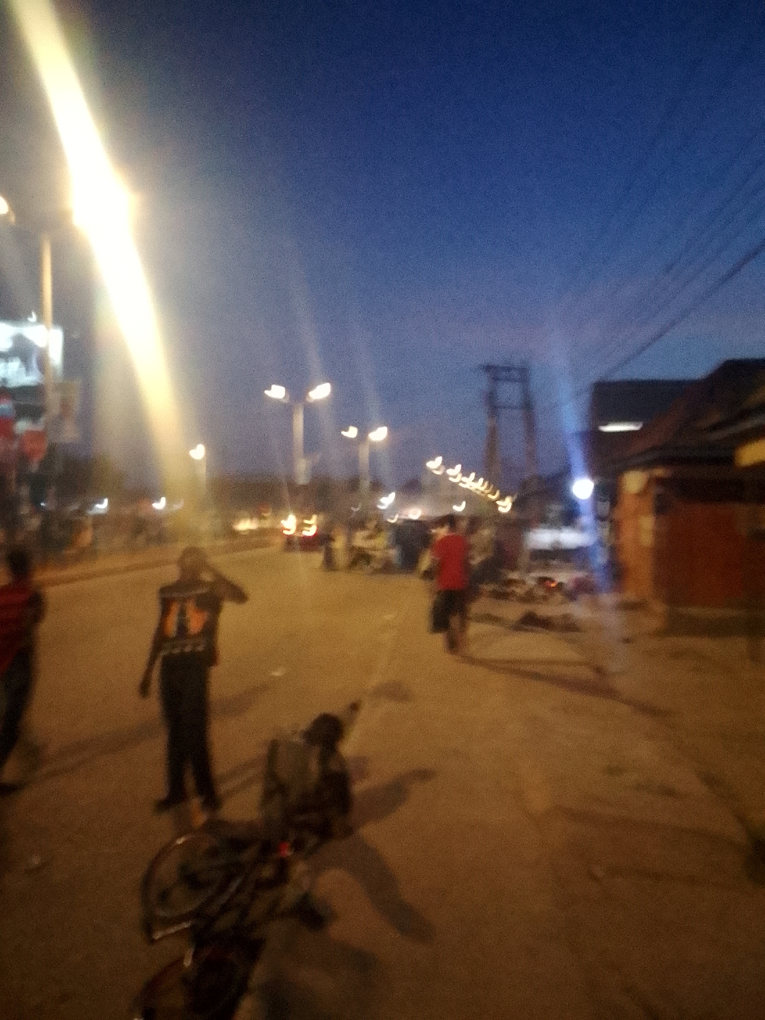 This media file presents a blurry, out-of-focus street scene at dusk or night in what appears to be an urban or semi-urban environment. The sky is a dark blue, indicating the late evening hours, with the primary illumination coming from bright artificial streetlights that produce prominent lens flares and light streaks across the image.
Several indistinct human figures are visible on a broad, light-colored ground surface, likely a road or open area. In the left foreground, a person wearing a red shirt is partially visible, moving away from the camera. More centrally, another figure stands, wearing a dark top with a lighter, illegible design on the back. Further back, multiple other figures are scattered along the street, their forms highly blurred.
A bicycle or motorcycle is seen lying on its side in the foreground, near the central figure. Along the sides of the street, there are vague outlines of buildings or structures, and overhead power lines or cables are faintly visible against the sky in the upper right. The ambient light suggests a bustling, everyday street setting.
The significant blur throughout the image makes it challenging to discern specific details, activities, or emotions. No legible text is visible on any signs, clothing, or objects. The scene captures a moment of street activity under artificial light, consistent with the provided context of Bukuru, Nigeria.