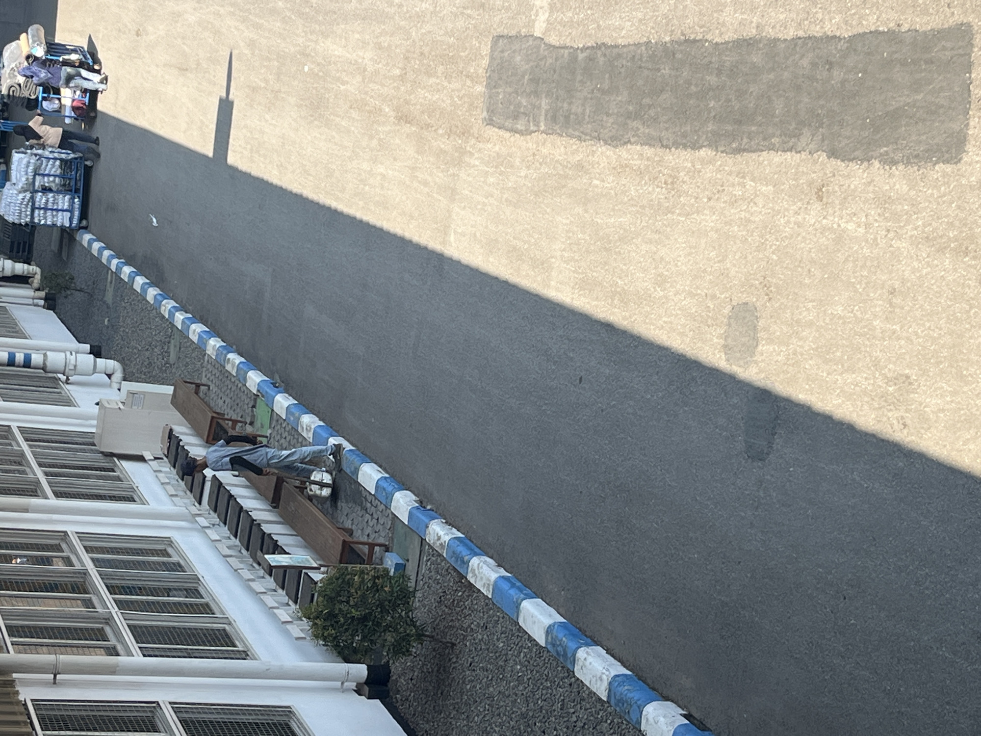 In the city of Pelang, Indonesia, a scene unfolds on a paved walkway bordering a building. The walkway is lined with a curb of alternating blue and white blocks. To the left of the walkway, a building with multiple windows is visible. A young man, dressed in a light-colored shirt and pants, stands on the walkway next to a bench. He appears to be holding a broom or similar cleaning tool. In the background, a cart loaded with white goods is being pushed by two individuals. Sunlight casts a long shadow across the paved area.