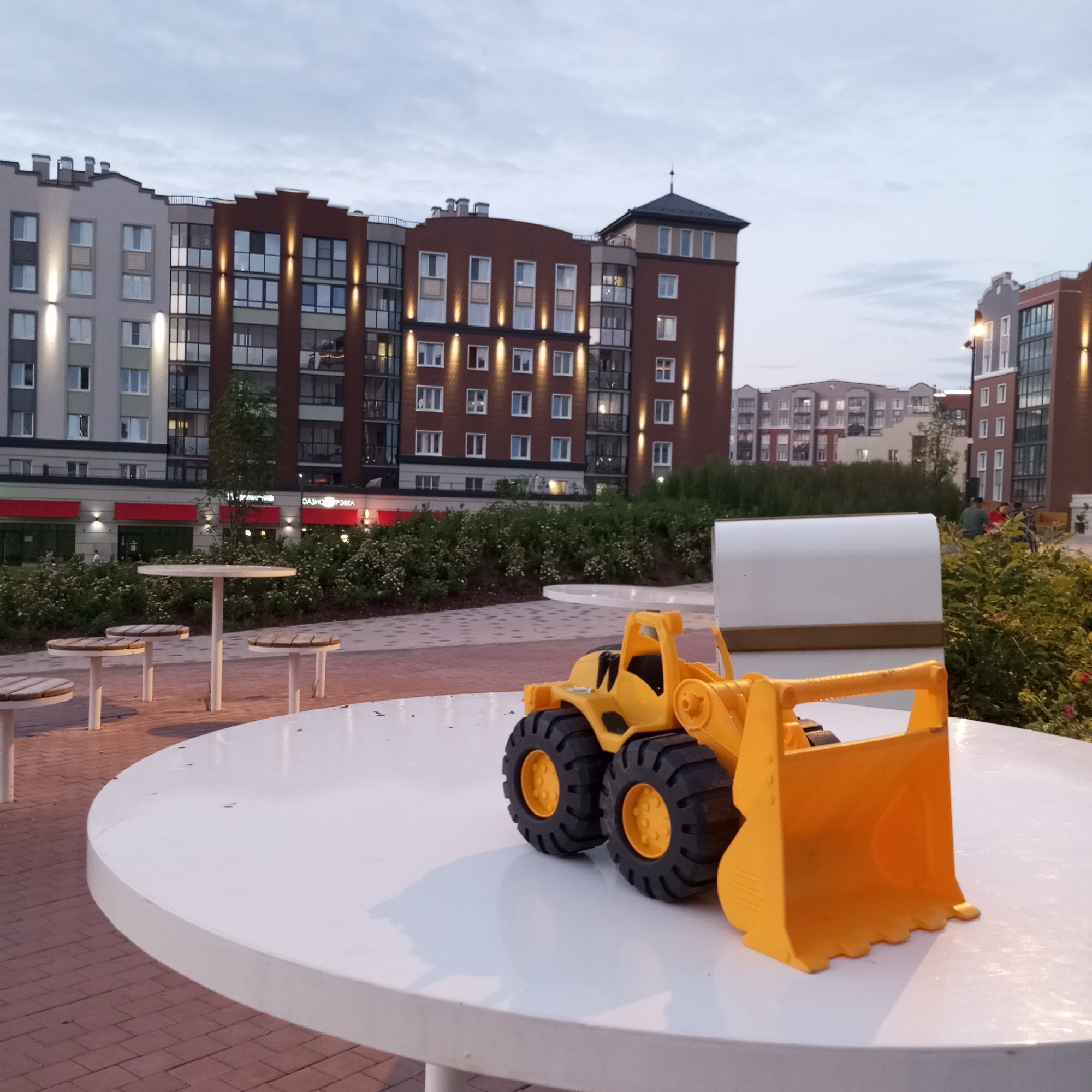 In the city of Pervomaysky, Russia, this image captures a toy yellow front-loader positioned on a large white circular table in an outdoor public space. Behind the table, a row of low-rise buildings with red awnings is visible, suggesting a commercial area. Further in the background, a cluster of multi-story apartment buildings with varying architectural styles and illuminated windows dominate the skyline under an overcast sky. The ground is paved with reddish-brown brickwork in a patterned arrangement. Small tables with wooden slat seats are scattered around the area, some with seating. Green bushes and trees provide a natural element to the urban landscape. The lighting in the scene indicates it is either early morning or late evening.