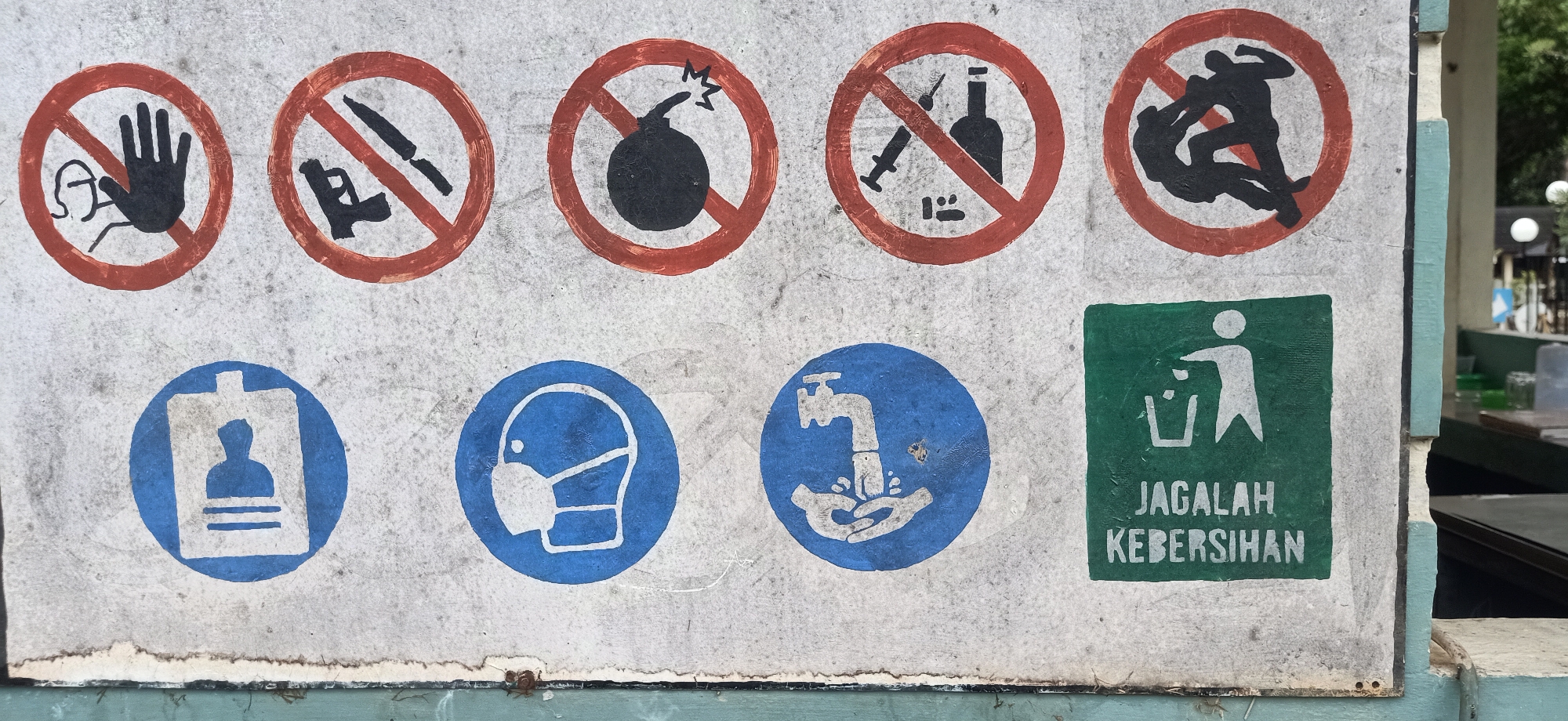 A weathered, multi-colored sign displays a series of pictograms and text, likely a set of rules or warnings, mounted on a textured, light-colored surface. The sign is positioned outdoors, adjacent to a teal-colored vertical beam.
In the top row, arranged from left to right, are five circular red symbols with diagonal red lines, indicating prohibition:
1. A black hand with an outline of a head behind it.
2. A black handgun and a knife.
3. A black bomb with jagged lines emanating from it.
4. A black syringe and a bottle.
5. Two black silhouetted figures in a wrestling or fighting pose.
In the bottom row, also from left to right, are three blue circular symbols and one green rectangular symbol:
1. A white silhouette of a person within an ID card holder.
2. A white silhouette of a person wearing a face mask.
3. A white depiction of a faucet with water flowing onto a pair of hands.
4. A green rectangle containing a white pictogram of a person throwing an item into a trash can, with the white text "JAGALAH" above "KEBERSIHAN" below, which translates to "MAINTAIN CLEANLINESS".
The sign shows signs of wear, with peeling edges and faded paint in some areas. The background of the sign is a rough, speckled grey. To the right of the sign, a blurred background suggests an outdoor environment with foliage and possibly some structures. The overall scene appears to be in Tambontunong, Indonesia.