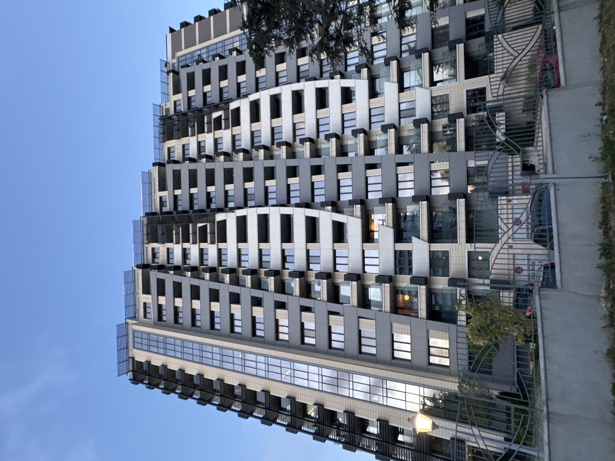 A low-angle view of a tall, modern apartment building in Varna, Bulgaria. The building has a repeating pattern of windows and grey cladding with occasional white curved architectural features. A dark tree partially obscures the view at the top. The building appears to be located in a residential area as there is a dark-colored sedan in the background and a metal fence. The sky is light blue and cloudless. A street lamp is visible behind the fence.