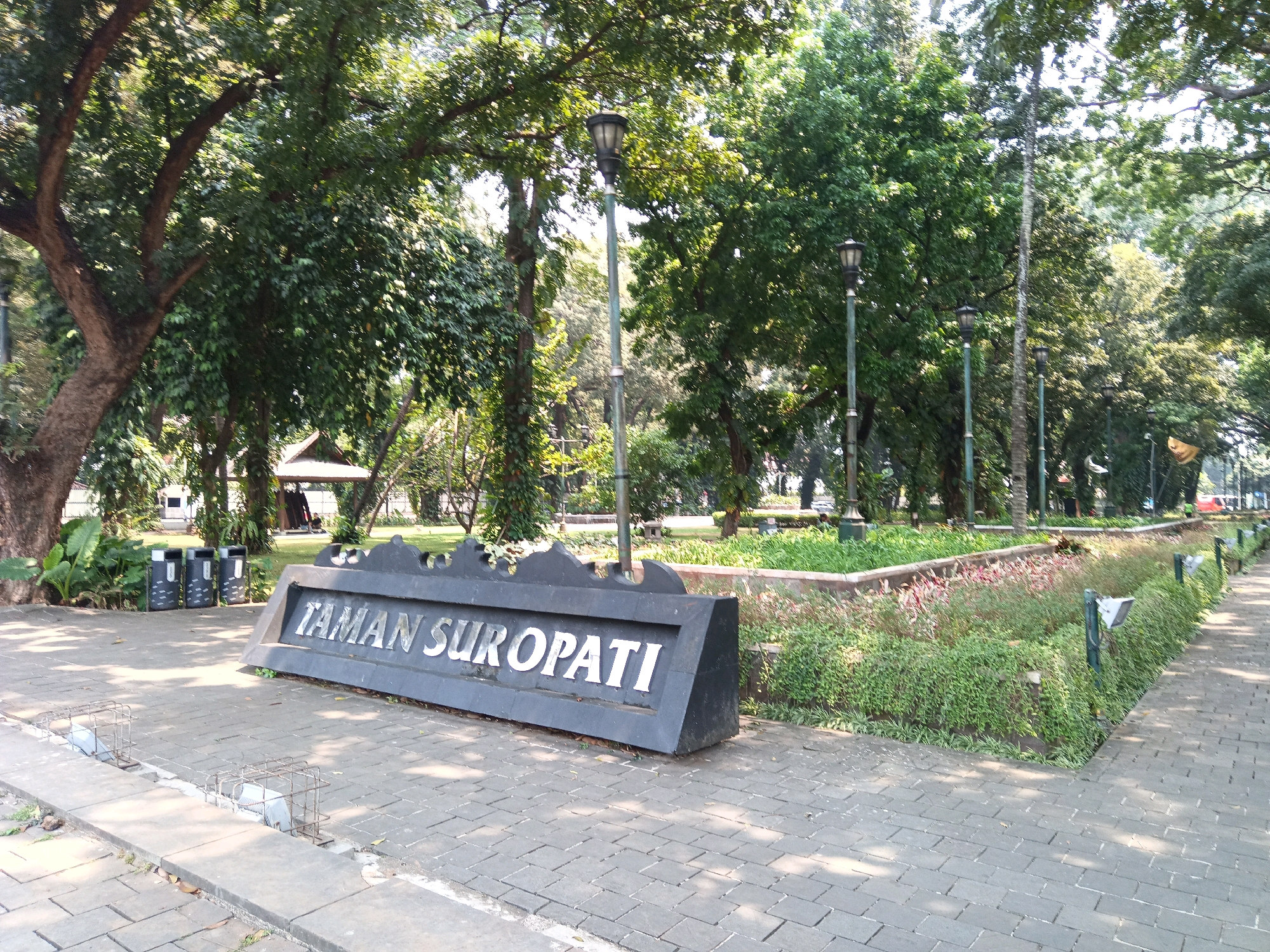 A paved pathway leads through a lush park setting with large trees and manicured greenery. In the foreground, a large, dark sign reads "TAMAN SUROPATI" in silver lettering. The park appears to be a public space, possibly a garden or memorial, with several lampposts strategically placed throughout. In the background, a small pavilion-like structure is visible amongst the trees, suggesting recreational or resting areas. The scene is bathed in natural sunlight, casting shadows on the pathway and highlighting the vibrant foliage. The overall atmosphere is serene and inviting, characteristic of a well-maintained urban park in Central Jakarta, Indonesia.