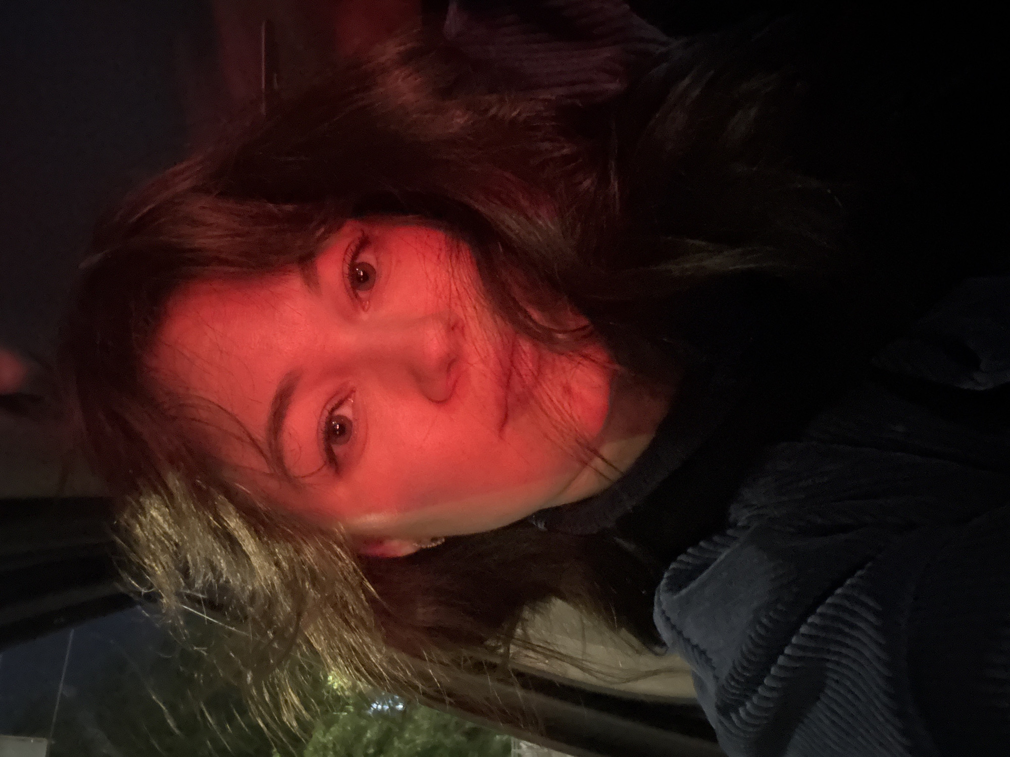 A young adult female is depicted in a close-up, dimly lit portrait, likely taken within a vehicle at night in Perm, Russia. Her face, positioned centrally, is significantly illuminated by a strong red light source from the viewer's left, casting the right side of her face and upper body into deep shadow. She has long, wavy dark brown hair, appearing somewhat disheveled, with strands falling across her right cheek and obscuring part of her mouth. Her eyes are open and directed towards the camera, revealing brown irises. A small, light-colored earring is visible in her left ear. She is wearing a dark blue corduroy garment, visible on her left shoulder and upper arm, with a distinct ribbed texture. A dark strap, consistent with a seatbelt, crosses her chest on the right side. The background is largely dark and indistinct, with faint ambient green light suggesting an exterior night scene visible through a window on the far left. Her expression is neutral or subtly contemplative.