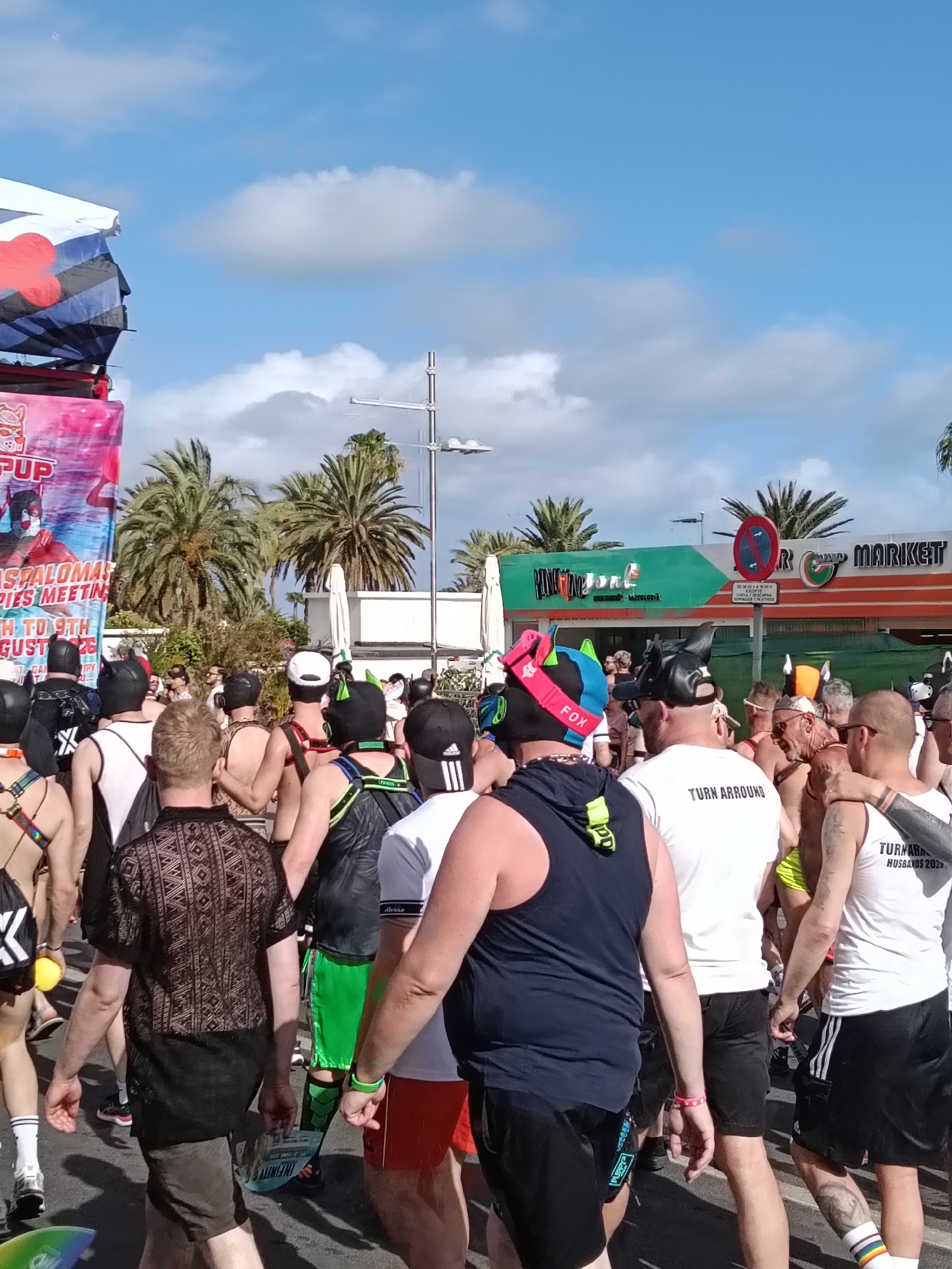 The image shows a crowd of people, many of whom are wearing fetish gear, particularly leather pup hoods, likely participating in a gay pride event. The scene is set in Playa Del Inglés, Spain, as evidenced by the "Superdune Market" signage and palm trees in the background. The event appears to be associated with the "Maspalomas PUPPIES MEETING," dates "4TH TO 9TH AUGUST 2023" visible on a banner to the left. The sky is partly cloudy with patches of blue. Many of the people are wearing shorts and t-shirts. One shirt reads "TURN ARROUND HUSBANDS 2023". Some are shirtless and wearing harnesses. There are various shops and structures visible in the background. A street sign is also visible.