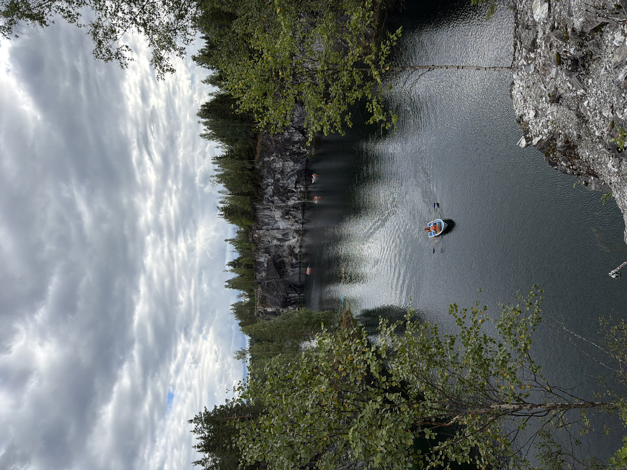 The image depicts Ruskeala Mountain Park in Ruskeala, Russia, a natural and historical park known for its marble canyons and emerald waters. In the foreground, a person wearing an orange life vest rows a small white boat with blue trim using oars. The boat is positioned on a dark, calm body of water, with ripples indicating movement. The water reflects the cloudy sky and the surrounding trees and cliffs.
In the middle ground, several other small boats with orange-vested occupants are visible on the water. The boats are scattered across the lake, some closer to the shore, others further out. The water is surrounded by steep, rocky cliffs, exhibiting a marbled texture of various shades of gray and white. Lush green trees and coniferous forests line the tops of these cliffs, extending into the background. The sky is overcast with heavy gray clouds, with a small patch of blue sky visible in the upper left quadrant. The overall atmosphere is serene and natural, with evidence of human activity in the form of recreational boating.