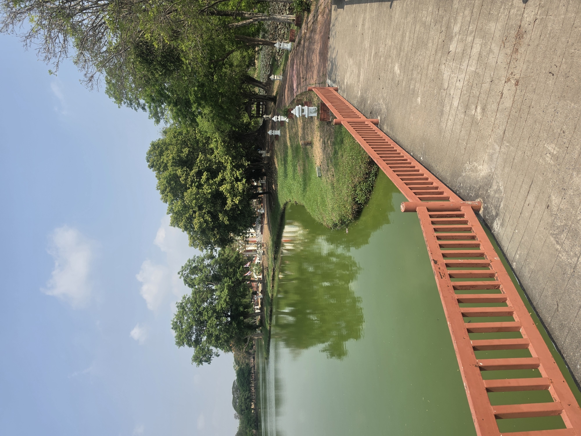 The image shows a scene in Phra Nakhon Si Ayutthaya City Municipality, Thailand, featuring a body of water alongside a concrete pathway. A red wooden structure, resembling a bridge or walkway, extends from the path over the water. The water has a green tint. Trees with lush green foliage line the side of the path opposite the water. The sky is light blue with scattered clouds. The image appears to be taken during daylight hours. Small white structures, possibly lanterns or decorative elements, are situated along the path.