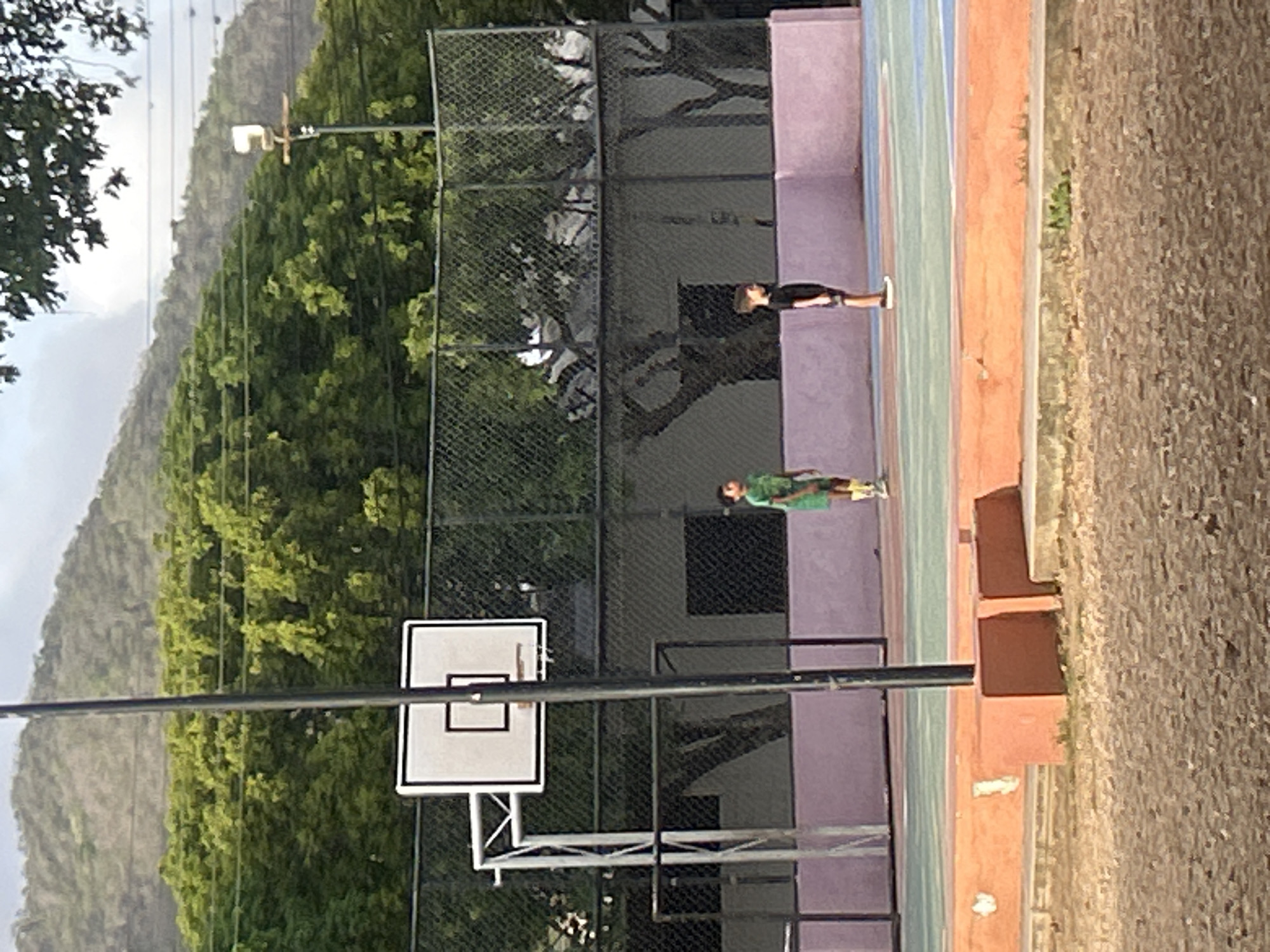 The image depicts an outdoor basketball court in Santa Ana, Venezuela. In the foreground, a dirt surface is visible, leading to the court. The court itself has blue and green sections, with an orange border. A chain-link fence encloses the court. A basketball hoop with a backboard is positioned on the left side of the frame. Two young boys are present on the court. One boy, wearing a green shirt and shorts, stands near the center. To his right, another boy in a black shirt and shorts stands facing left. In the background, lush green trees and a sloping hillside under a hazy sky suggest a natural landscape surrounding the urban setting.