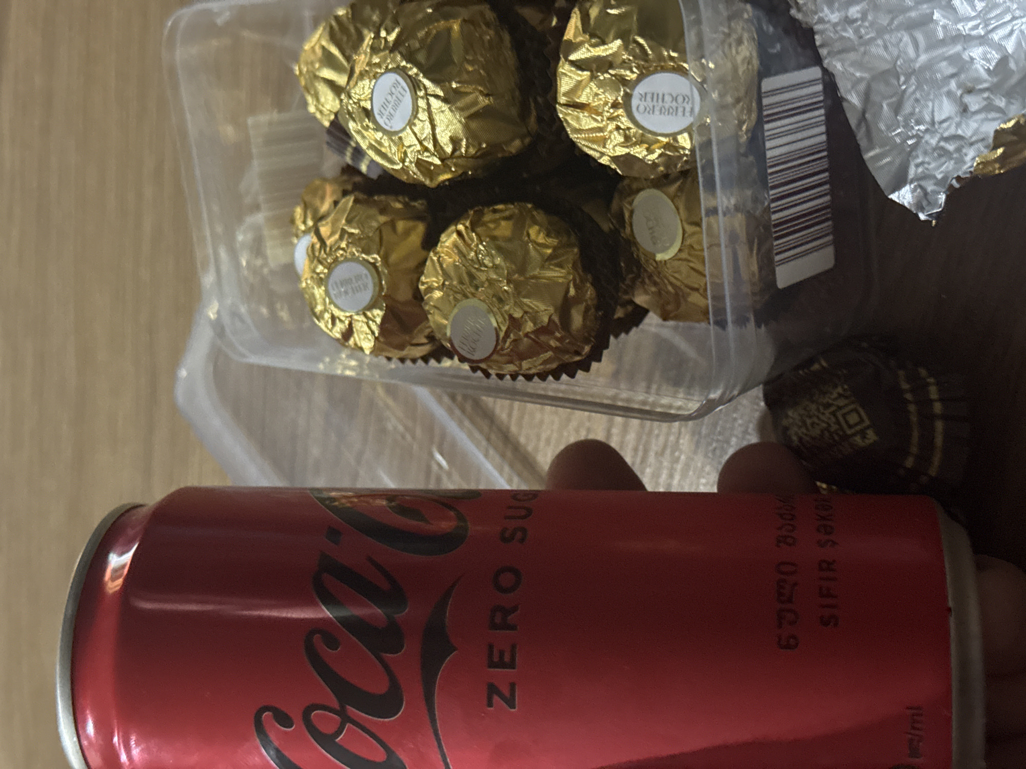 The image shows a Coca-Cola Zero Sugar can and a container of Ferrero Rocher chocolates on a wooden surface. The Coca-Cola can is red with the Coca-Cola logo and "ZERO SUGAR" printed on it. There are some inscriptions in Georgian script below "ZERO SUGAR". The Ferrero Rocher chocolates are individually wrapped in gold foil and are in a clear plastic container. Part of a barcode is visible on the container. The scene appears to be indoors, possibly in a home or office environment, with no specific identifying landmarks for Tbilisi, Georgia. The lighting suggests a natural light source from the side. The image is taken from a slightly elevated angle, focusing on the can and chocolates.