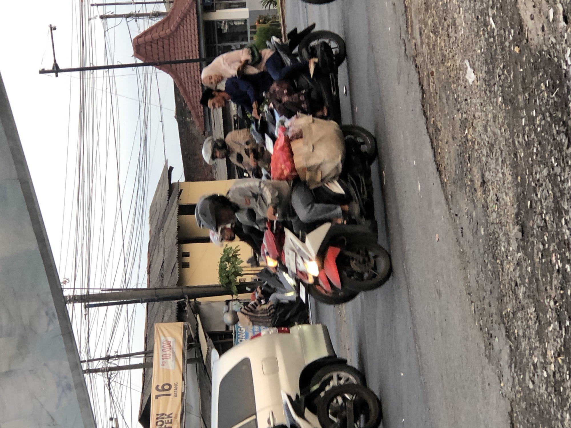 The image captures a street scene in Kludan, Indonesia, characterized by a moderately busy road with several motorbikes. In the foreground, a red and white motorbike is prominently featured, with a driver wearing a helmet and a backpack. Behind this, another motorbike carries three people: a man driving, a woman in a pink hijab as a passenger, and another individual behind her. Further to the left, a white van is partially visible, suggesting general traffic. The background shows a building with a distinctive red, multi-tiered roof, likely a shop or residence. Overhead, a dense network of electrical wires crisscrosses the sky, indicating a developed urban or semi-urban area. A banner with text and numbers, possibly advertising, is visible on a building to the left. The road surface appears asphalted but with some unevenness and debris. The overall lighting suggests it is daytime, possibly late afternoon, with overcast skies.