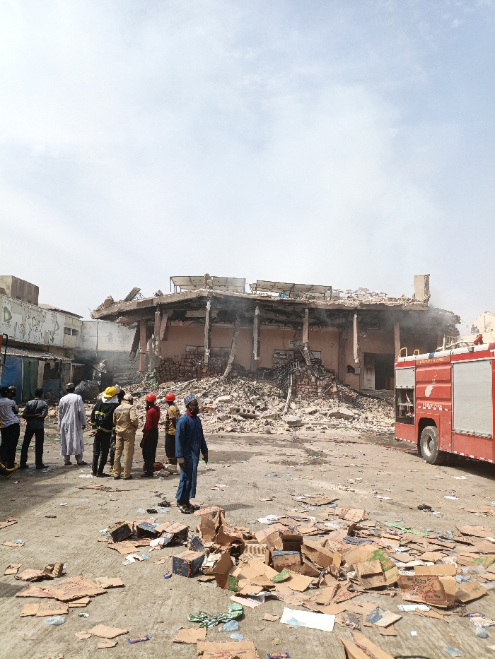 The scene in Fagge C, Nigeria, depicts the aftermath of a structural collapse and fire impacting a multi-story building. The structure shows severe damage, with upper floors partially caved in, exposing charred concrete columns and beams, and extensive rubble accumulated at its base. Active smoke plumes are visible emanating from the wreckage. A red fire truck is partially visible, positioned on the right side of the frame. Approximately twelve individuals are present, observing the damage. Five individuals are identified as firefighters, wearing protective gear and helmets (red and yellow), while the remaining individuals are in civilian attire. The immediate foreground ground plane is covered with scattered cardboard boxes and miscellaneous debris.
