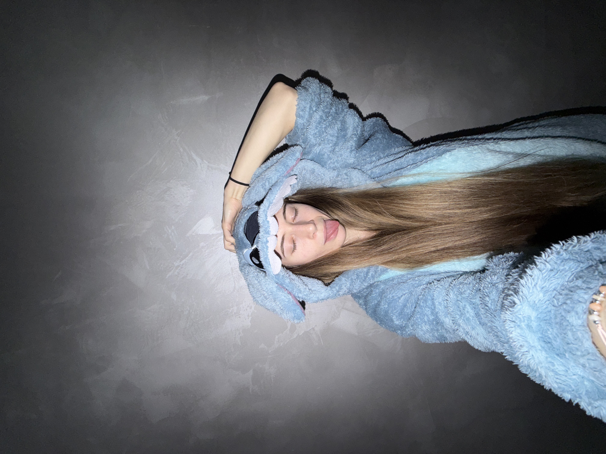 A person wearing a blue "Stitch" onesie lies supine against a gray, textured wall. The person's hair is brown and long. The person's eyes are closed and tongue sticks out. A black hair tie is visible on the person's left wrist. The photo's origin in Plovdiv, Bulgaria is undetermined.