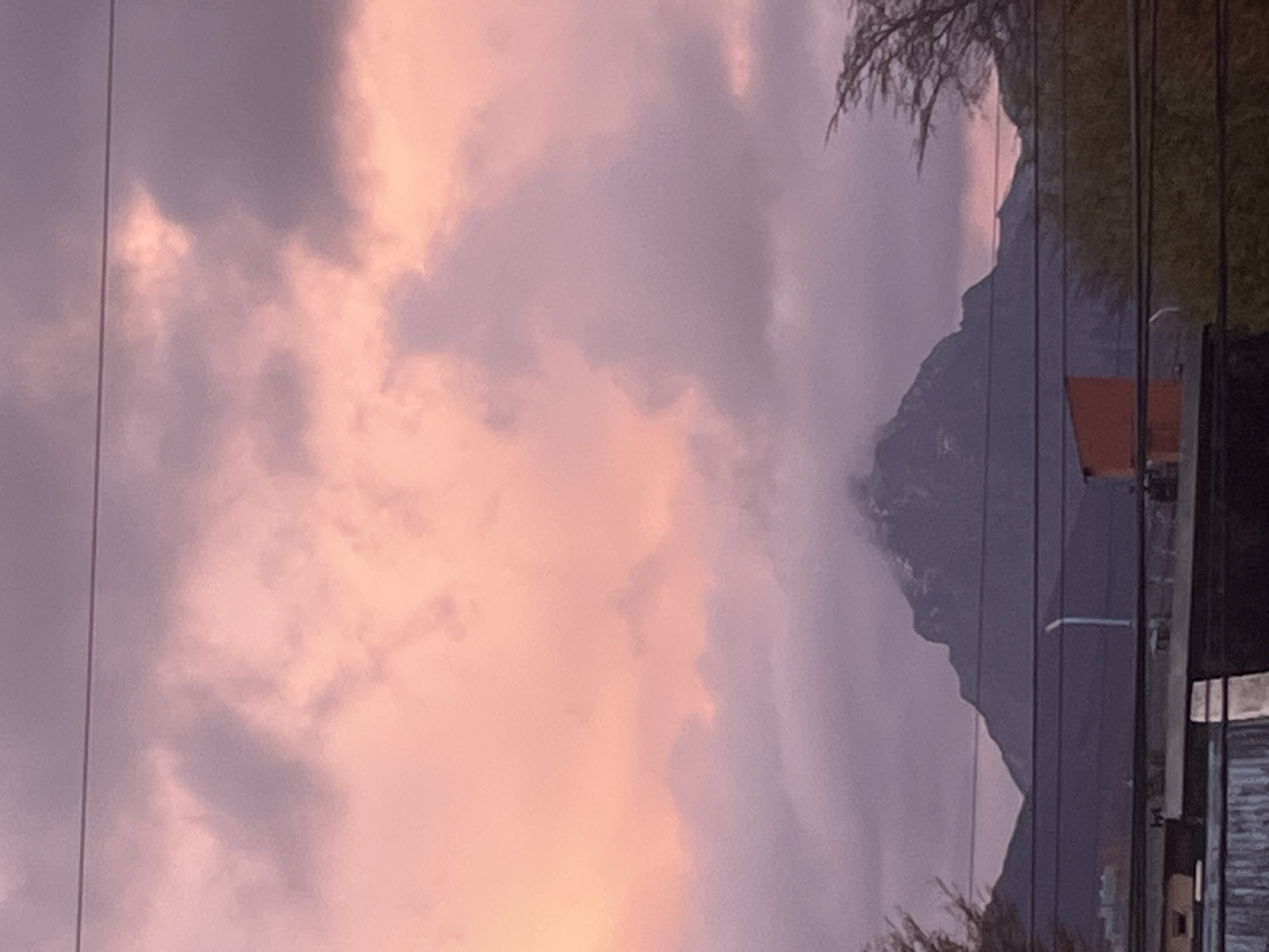 The image captures a serene twilight scene, likely either sunrise or sunset, dominated by a dramatic sky and a prominent mountain range. The sky is filled with soft, diffused clouds displaying a spectrum of pink, orange, and purple hues, indicating the early morning or late evening light. Darker grey and purple clouds are interspersed with brighter, more vibrant patches.
In the background, a large, dark mountain range is silhouetted against the colorful sky, its rugged peaks defining a strong horizon line. The mountain appears imposing and grand. Several thin, dark lines, likely power lines or utility cables, traverse the frame vertically and horizontally, cutting across both the sky and the mountain.
To the right side of the frame, subtle indications of human presence are visible: a small, rectangular orange structure and a white street lamp pole are discernible against the mountain's silhouette. Dark, indistinct vegetation, possibly trees or bushes, frames parts of the scene in the top and bottom right corners.
The scene is tranquil, devoid of any discernible activity or people. The cloud cover is significant, but allows the warm and cool tones of the twilight sky to glow through. The visible elements suggest a location where nature and some human infrastructure coexist, consistent with the mountainous and somewhat remote setting of Ushuaia, Argentina. No visible text is present.