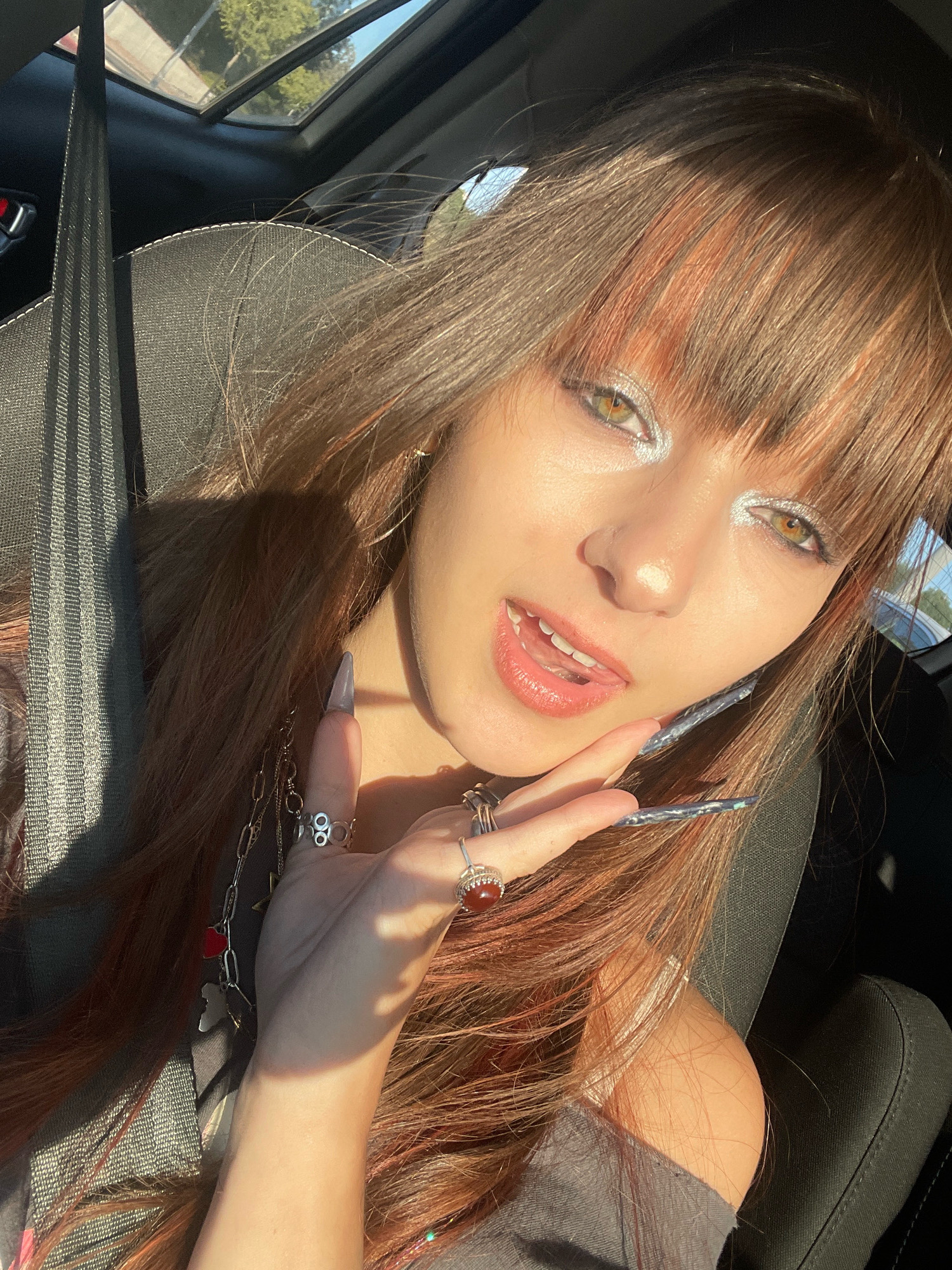 A young woman with long brown hair, some with reddish highlights, and bangs is sitting in the passenger seat of a car. She is wearing silver eyeshadow and has long, sharp, dark nails. She is wearing rings on her fingers and a chain necklace with charms. Her mouth is open, and her hand is resting on her cheek. The sun is shining through the car's sunroof, creating shadows. The car's interior is visible, including a seatbelt and dark upholstery. The exterior of the car shows a blurry view of trees and sky.