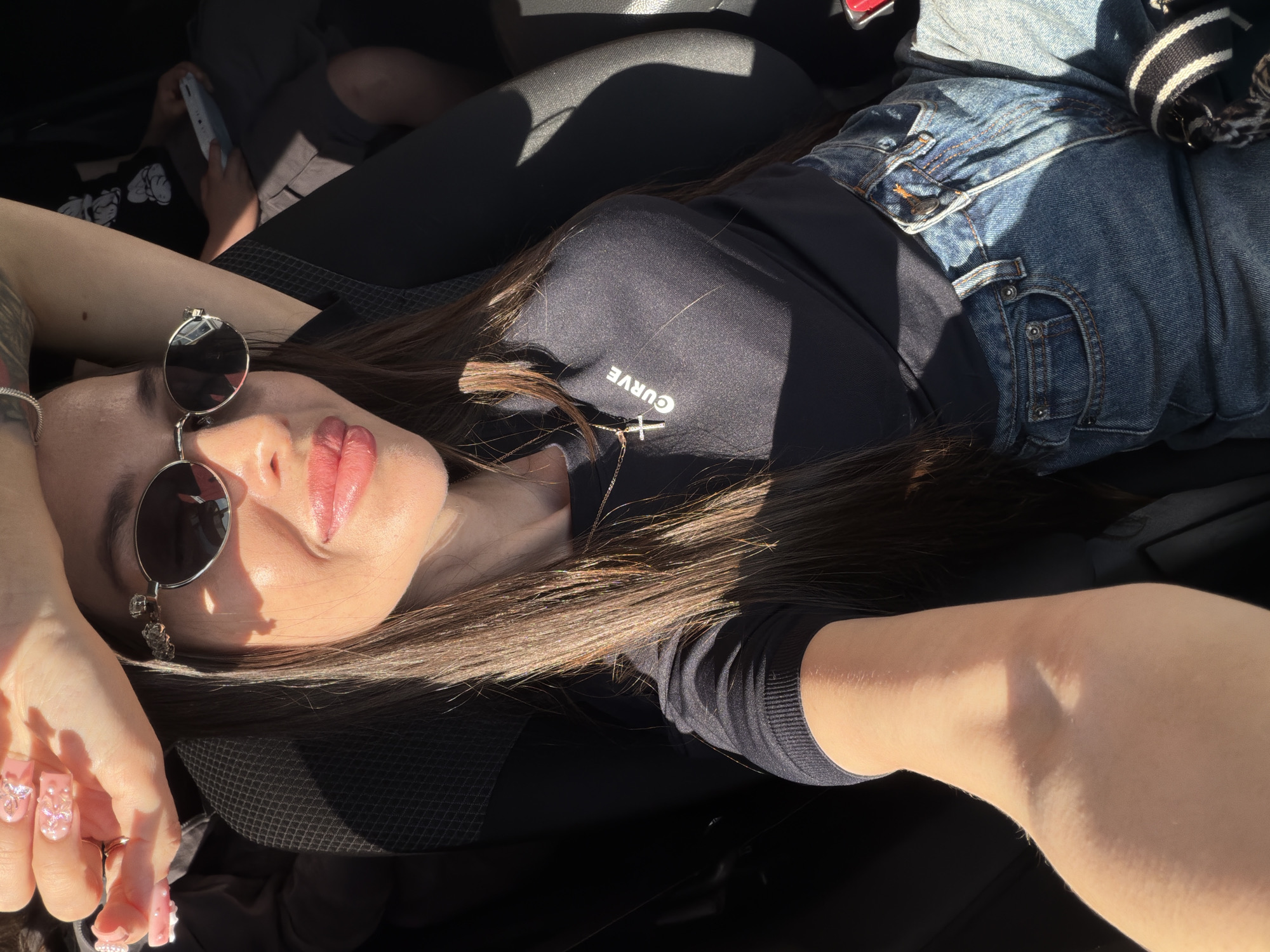 A high-angle selfie captures a woman reclining in the sunny interior of a car, with a second person partially visible in the background. The primary subject lies across the vehicle's seats, her long, dark hair spread out. She wears round, dark sunglasses, a black long-sleeve top with the word "CURVE" printed upside down on the chest, and a gold cross pendant necklace. Her lower body is clad in blue denim jeans worn over black leggings. Her lips are a glossy pink, and her long artificial nails are pink with glitter and embellishments. Her expression is relaxed, with her eyes hidden behind the sunglasses, suggesting she is enjoying the bright, direct sunlight streaming into the vehicle.
In the background, a person, possibly a child, is visible in grey shorts and a black t-shirt with a white graphic, engrossed in a white smartphone. The strong sunlight creates clear highlights and deep shadows throughout the scene, emphasizing the daytime setting. The woman appears to be holding the camera for the selfie with her right hand, which is visible in the foreground. The overall scene conveys a casual, relaxed moment inside a car on a bright day in Izhevsk, Russia.