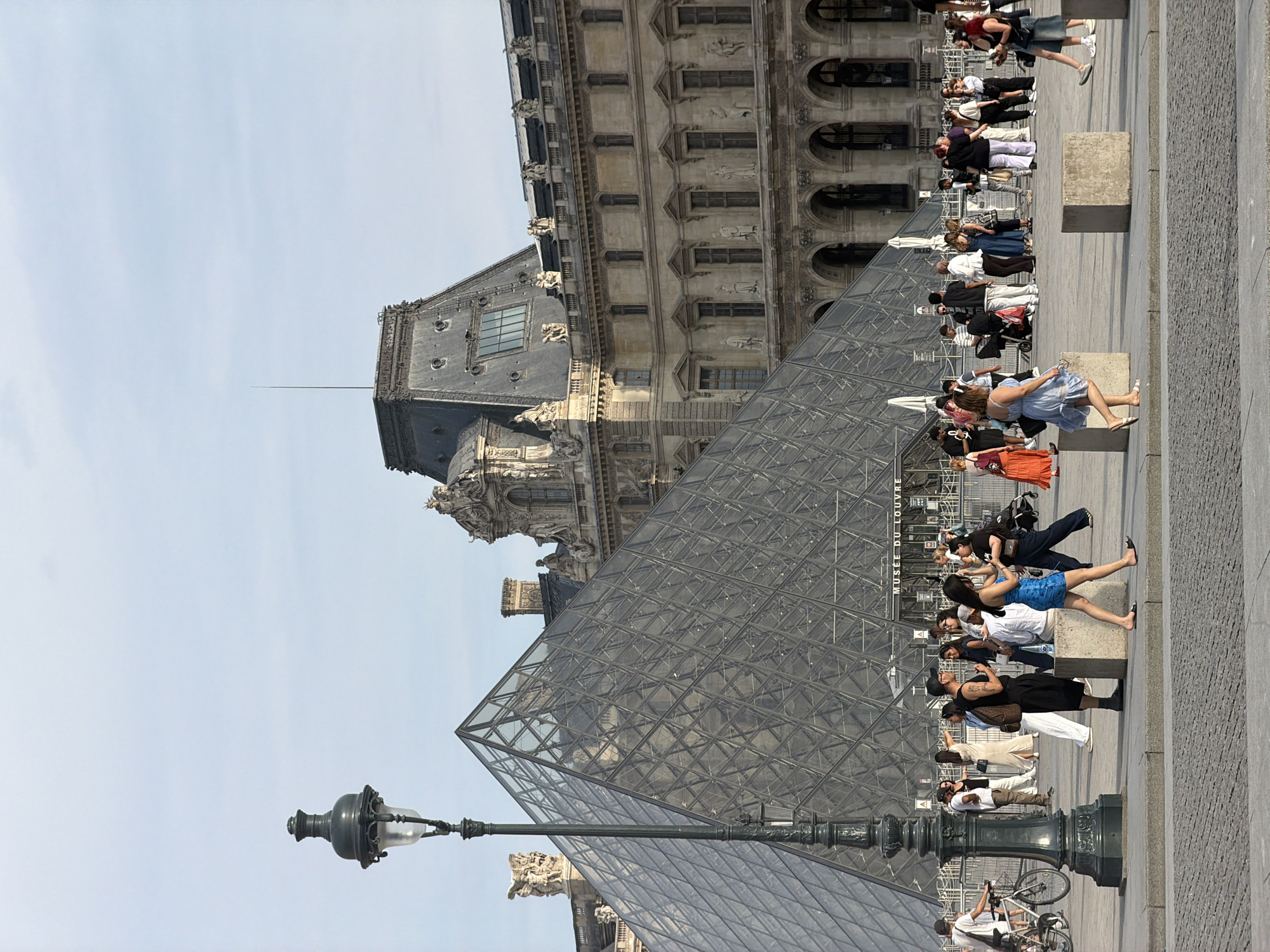 The image depicts a daytime scene outside the Louvre Museum in Paris, France. The iconic glass pyramid of the Louvre is prominently featured on the left side of the frame, reflecting the sky and surrounding architecture. To the right of the pyramid is the grand facade of the Louvre building, characterized by its classical architectural style, with numerous windows and arched entrances.
In the foreground and midground, a diverse group of people are seen walking, standing, and interacting. Many appear to be tourists, with some carrying bags and cameras. A street lamp stands tall in the left foreground. The ground is paved with cobblestones in the lower portion of the image, transitioning to a smoother, lighter-colored surface in the midground where people are gathered. The sky is a pale blue with scattered clouds, suggesting fair weather. The overall impression is of a bustling public space on a pleasant day.