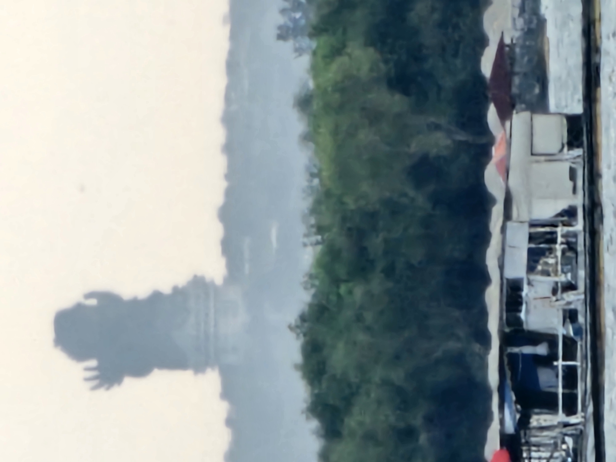The image depicts a hazy, distant view of the GWK Cultural Park in Denpasar, Indonesia. In the background, silhouetted against a pale sky, stands the iconic Garuda Wisnu Kencana statue, appearing as a large, dark figure with raised hands. Below the statue, a dense line of green trees forms a horizontal band across the middle of the frame. In the foreground, a body of water is visible, with a floating structure, possibly a boat or a platform, partially obscuring the view of the shore. This structure has a makeshift appearance with several sections covered by roofs of various colors, including red and white. Piles of what appear to be sand or building materials are visible on the shore behind the floating structure, along with a small, dark red-roofed building. The overall impression is one of a somewhat hazy and distant landscape, with the monumental statue dominating the background.