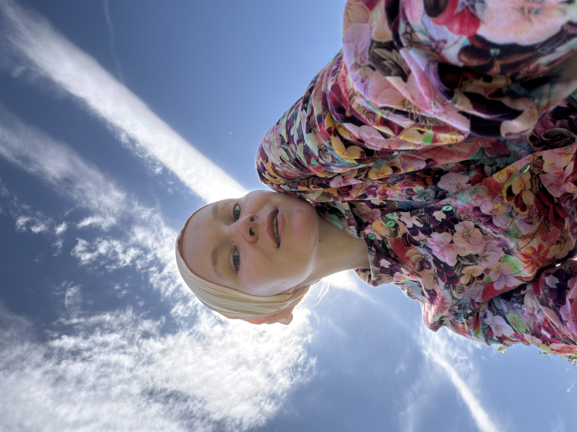 A woman with light skin, light eyes, and a peach-colored head covering looks down at the camera from a low angle. She is wearing a floral-patterned shirt with various colors including pink, red, yellow, and green. The background is a bright blue sky with streaks of white clouds. The location is presumed to be Kadryakovo, Russia, based on the user prompt.