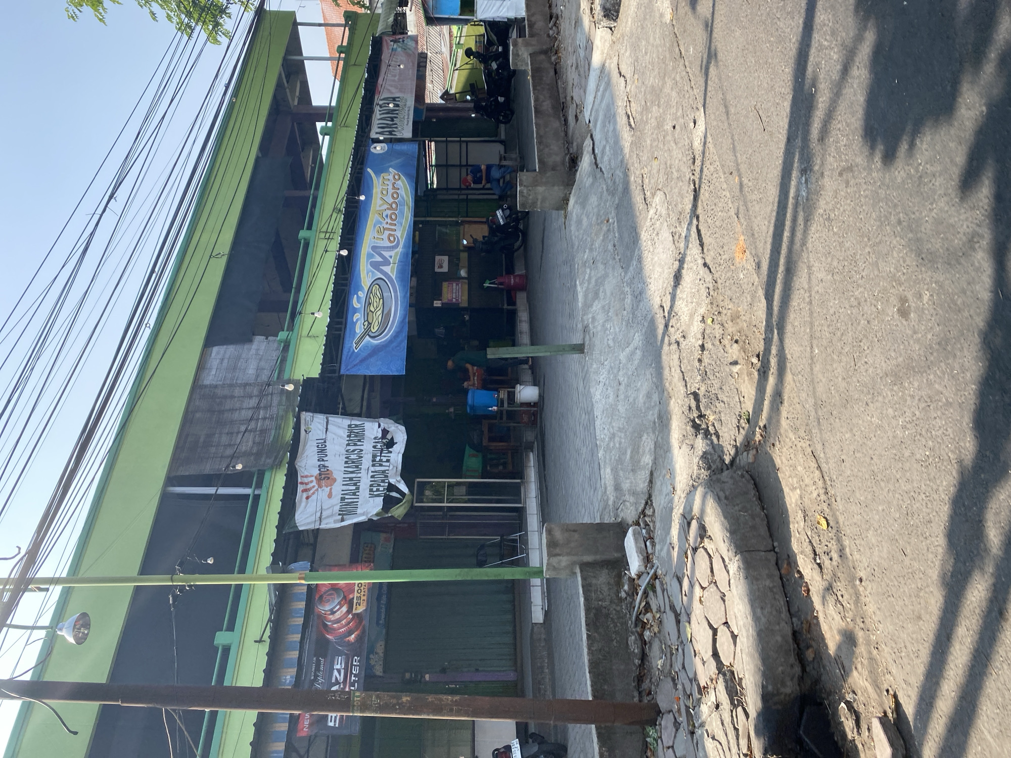 The image depicts a street scene in Kediri City, Indonesia, likely in front of a row of shops or food stalls. A bright green, two-story building with a black awning dominates the background, with numerous electrical wires crisscrossing overhead. In the foreground, a paved walkway with a concrete curb separates the street from the building.
Several shops are visible along the building's ground floor. One shop has a sign that reads "Mie Ayam Malioboro," indicating it's a noodle restaurant. Another banner on the building displays a red handprint and text in Indonesian that translates to "Stop Illegal Levies - Ask for Parking Tickets from Officials."
Various objects are present: parked motorcycles, chairs, tables, a blue water container, and what appears to be a trash can. Several people are visible, including one person sitting on a chair outside a shop, wearing a red cap. Another person is standing near a table, and a third person is partially visible behind a stall.
The street itself is paved with asphalt, showing signs of wear and tear, with some cracked sections and debris. The overall impression is a lively, slightly cluttered urban environment. The lighting suggests it is daytime, with shadows cast by the building and overhead wires.