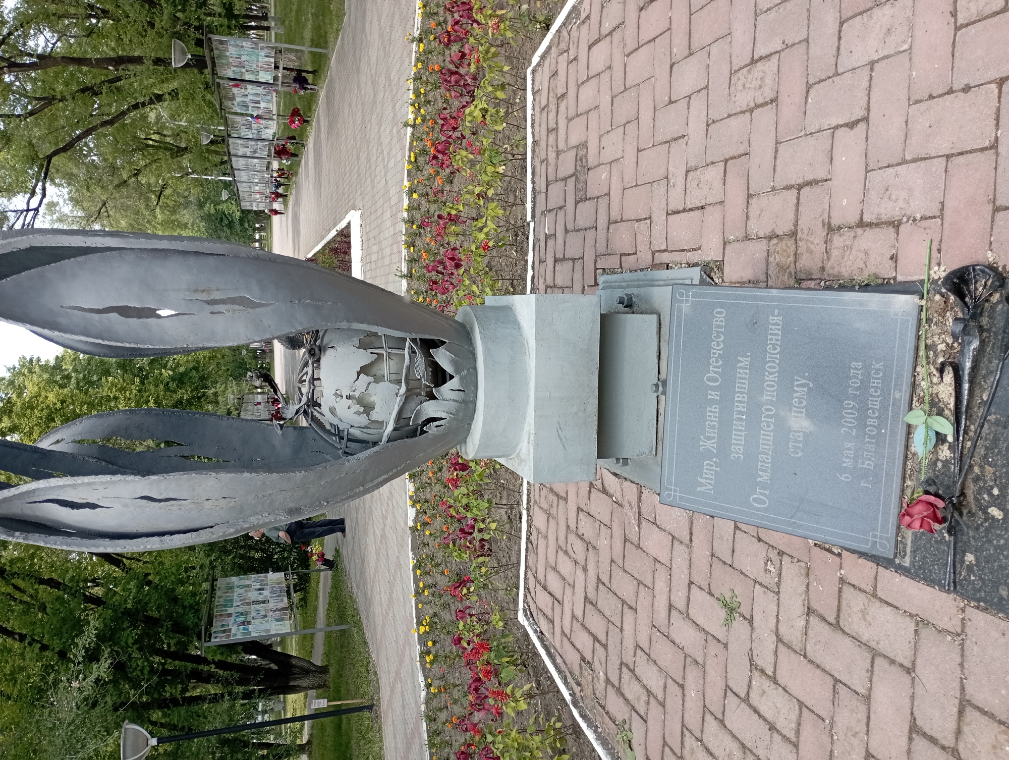 The image depicts a monument in the city of Blagoveshchensk, Russia. The monument features a large, abstract metal sculpture that resembles outstretched petals or flames, cradling a globe. In front of the sculpture is a stone plaque with Russian text. The area around the monument is paved with reddish bricks, and there are flower beds with red and yellow flowers. In the background, a pathway is lined with numerous display boards showcasing portraits, likely of people honored by the monument. A few individuals are visible in the distance, looking at the displays. The environment appears to be an outdoor public space, possibly a park or memorial area, with trees and lampposts.
