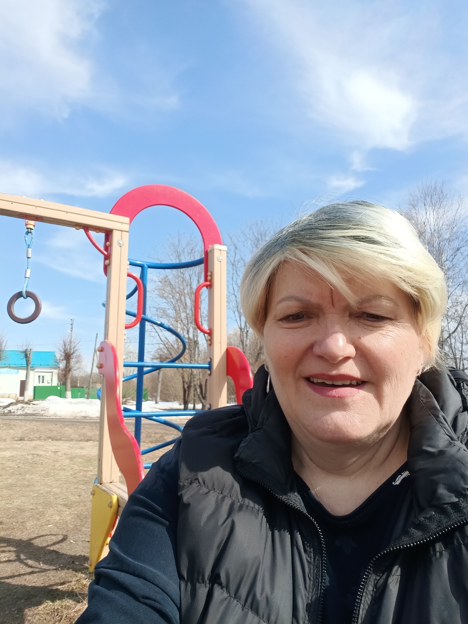 A woman is taking a selfie on a playground in Красный Маяк, Russia. The woman has short, blonde hair and is wearing a black vest. Behind her, there is a playground structure with red and blue components. In the background, there is a building with a blue roof, some melting snow, and bare trees. The sky is blue with some clouds.