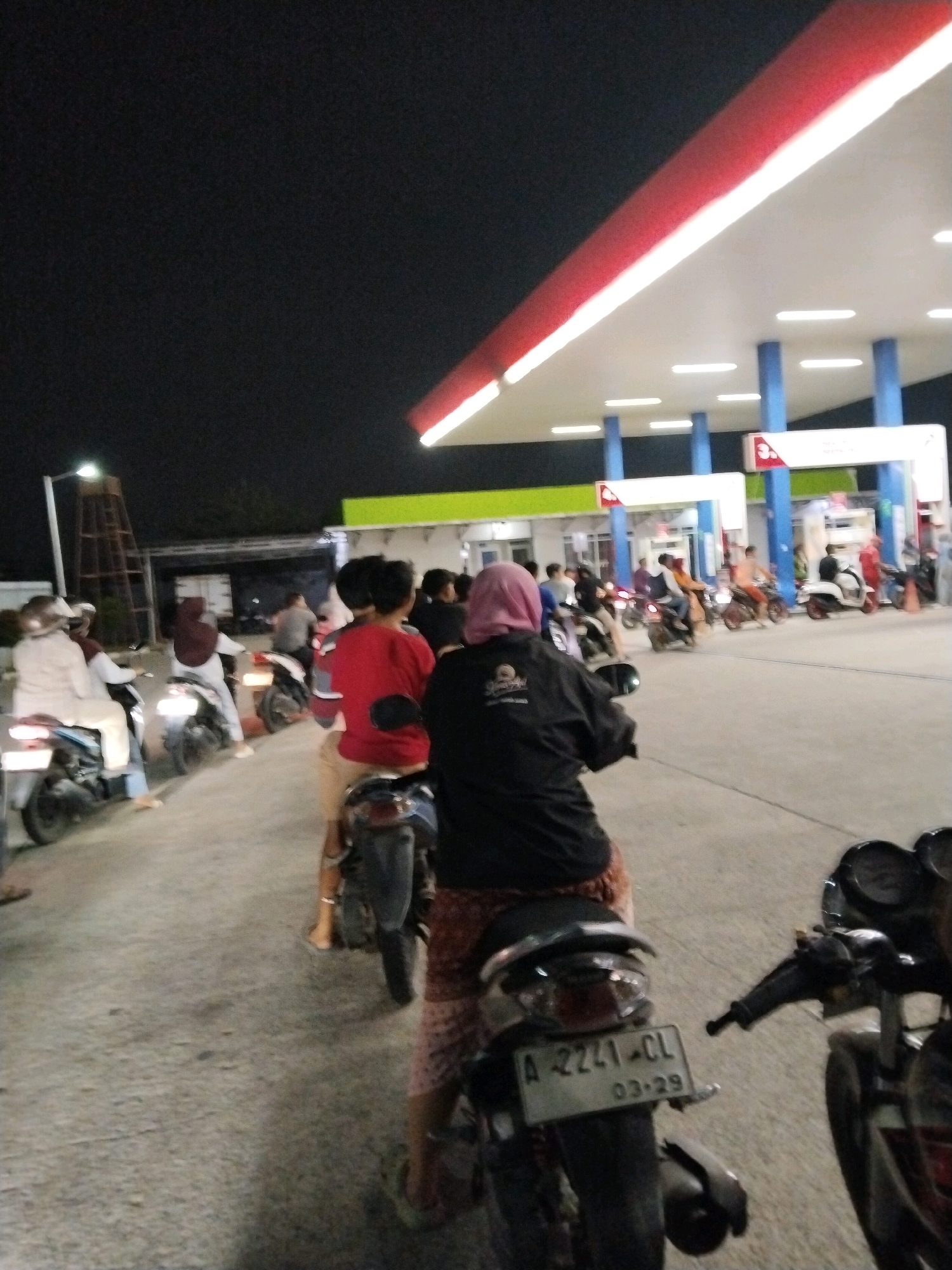In Serang, Indonesia, a nighttime scene unfolds at a brightly lit gas station. Multiple individuals on motorcycles are queuing, waiting for service. The gas station features a distinctive red and white canopy supported by blue pillars. Some individuals are wearing helmets and casual clothing, with one person in the foreground wearing a pink hijab and a black jacket. The ground is paved, and the surrounding environment is dark, illuminated by the artificial lights of the gas station and streetlights. One motorcycle in the foreground displays a license plate with the characters "A-2241-CL" and "03-29". The overall atmosphere suggests a routine activity of refueling vehicles during the evening.
