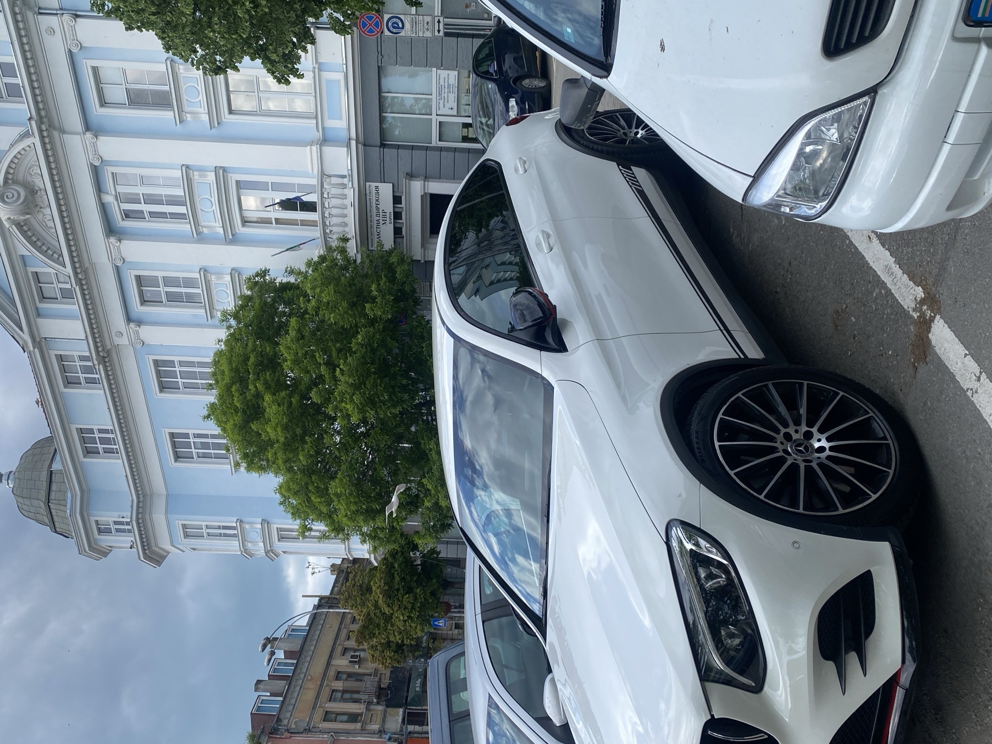 The image depicts a street scene in Varna, Bulgaria. The most prominent feature is a modern white Mercedes-Benz car parked on the street. The car appears to be a newer model with distinctive black accents along the side and on the wheels. Other vehicles are partially visible nearby, suggesting a busy urban environment.
In the background, there is a distinctive blue and white building with ornate architectural details. A tree partially obscures the view of the building. Further in the background, other buildings of various architectural styles are visible. Street signs are affixed to the building. The sky is overcast, which indicates either early morning or late afternoon lighting conditions.