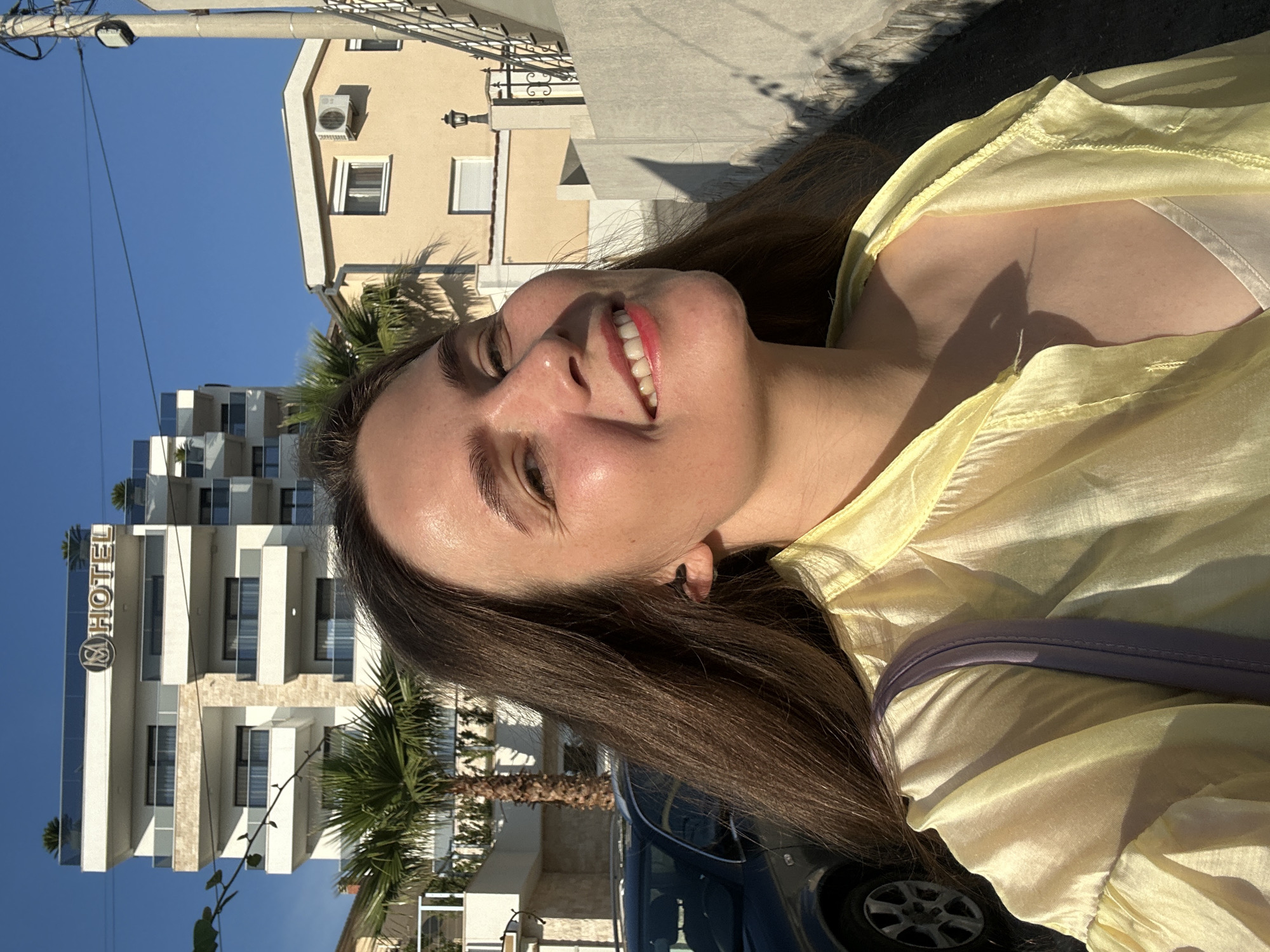 In Šušanj, Montenegro, a young woman with long brown hair smiles at the camera, her head tilted slightly to the right. She wears a light yellow, translucent shirt over a white top and a purple strap from a bag is visible across her shoulder. Behind her, a modern, white hotel building with multiple balconies and glass railings stands against a clear blue sky. A palm tree is positioned in front of the hotel. To the right of the hotel, a smaller, light-colored building with a dark-tiled roof is visible, featuring an air conditioning unit on its facade. A black car is parked on the left side of the image. A concrete wall runs along the right side of the frame, casting a shadow.