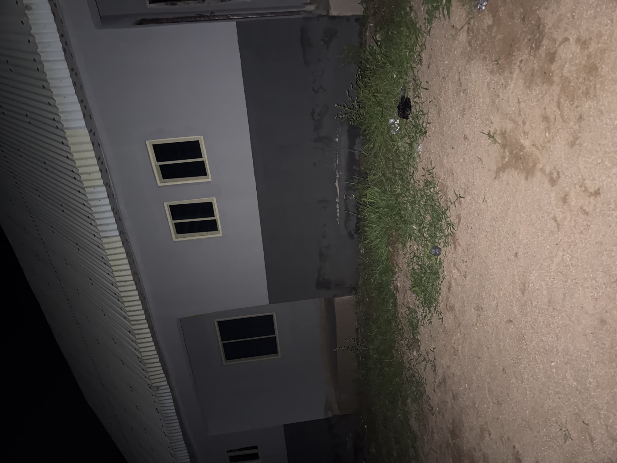 The image shows a plain, light grey building with a corrugated metal roof at night in Jauro Yinu, Nigeria. The building has two windows visible on the upper half of the wall, each with dark panes and light-colored frames. Below these windows, the building is painted a dark grey. The ground in front of the building is a mix of sandy soil and patchy grass. Some litter, including a dark, crumpled item, is visible in the grass area. The scene appears to be an exterior view of a residential or commercial structure in a somewhat unkempt environment.