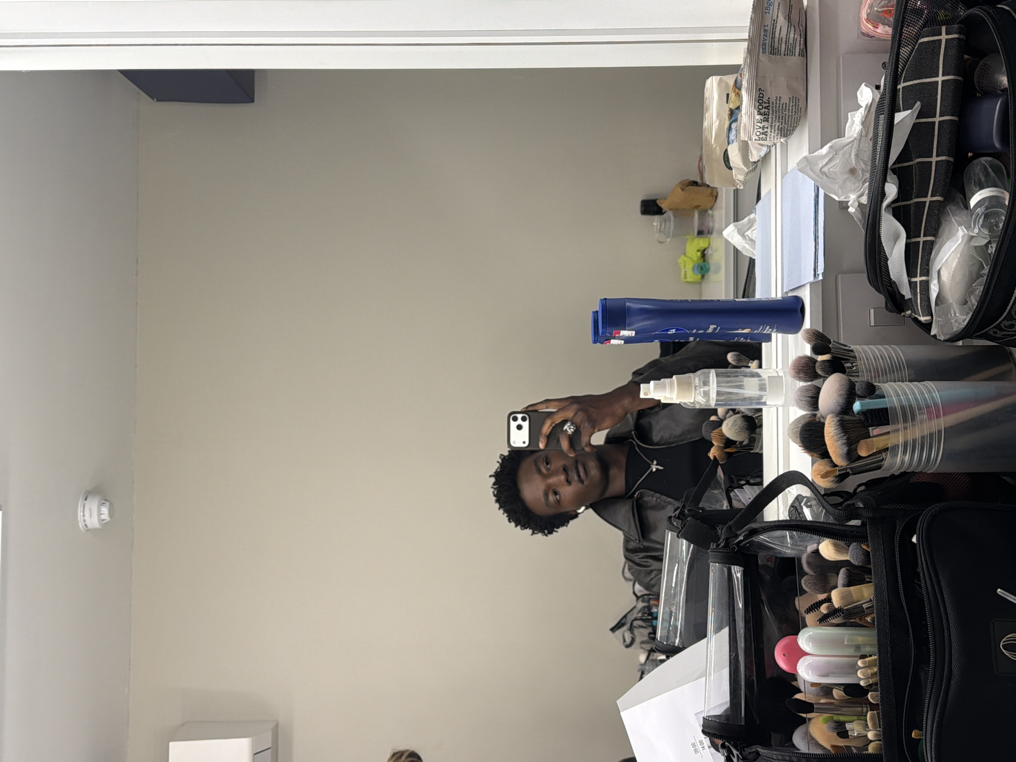 The image shows a reflection of a young man in a mirror. He is holding a smartphone and appears to be taking a selfie. He is wearing a black leather jacket and a necklace with a cross pendant. His hair is short and curly.
To the right of the man in the reflection, on the surface reflected in the mirror, is a makeup artist's setup. This includes various makeup brushes in containers, a clear bottle with a spray nozzle, a blue tube, bags of snacks, makeup wipes, and several makeup bags.
In the background, the wall is painted a light beige or off-white color. A smoke detector is visible on the left side of the wall, and a small white object is positioned below the smoke detector. A dark rectangular object is attached to the upper section of the wall.
It is not possible to determine the location in the city of Manchester, United Kingdom, from the given image.
