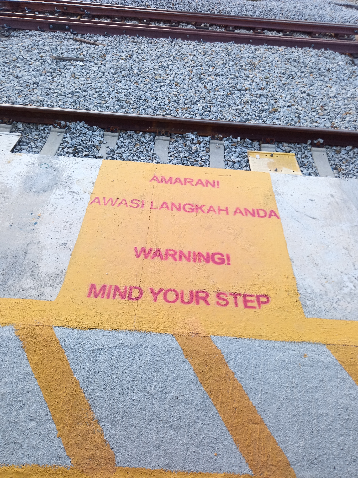 A close-up, overhead shot captures a textured concrete surface painted with yellow and pink markings at a railway station in Klang City, Malaysia. Prominently displayed in pink stencil lettering are cautionary messages. The top line reads "AMARAN!", followed by "AWASI LANGKAH ANDA" in Malay, translating to "Watch your step." Below this, in English, are the phrases "WARNING!" and "MIND YOUR STEP." The yellow paint is applied to a raised section of concrete, likely a platform edge, with diagonal yellow and grey stripes painted on the surrounding grey concrete, indicating a hazard or crossing area. In the background, out of focus, are gravel ballast and parallel railway tracks.