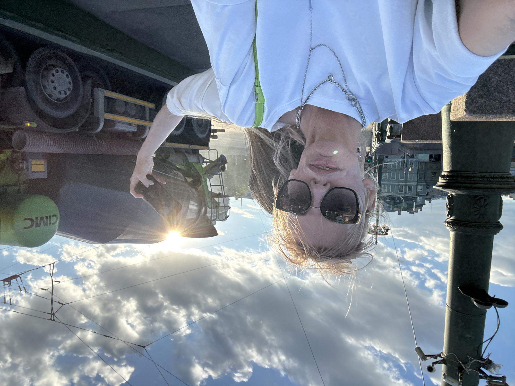 A woman with blonde hair and sunglasses takes a selfie outdoors in Saint Petersburg, Russia, with a cement truck in the background. She is wearing a white t-shirt and a silver necklace. The woman's left arm is extended, holding a dark object, possibly a phone or camera, up towards the cement truck. The truck has a green CIMC logo on its side. The sun is visible, casting a bright glare. In the background, there are buildings and a cloudy sky. The scene appears to be on a bridge, indicated by the railing and structural elements.