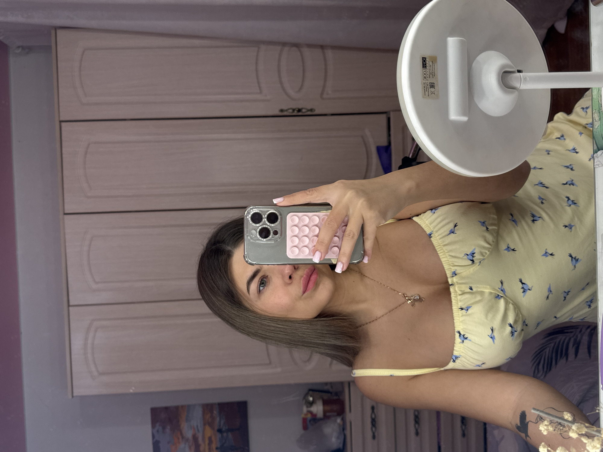 A woman is taking a selfie in front of a mirror. She is holding a phone in front of her face. She is wearing a yellow dress with blue bird patterns. She has a necklace and a tattoo on her arm. Behind her is a wardrobe and a ring light. The location is suggested to be Chaikovsky, Russia.