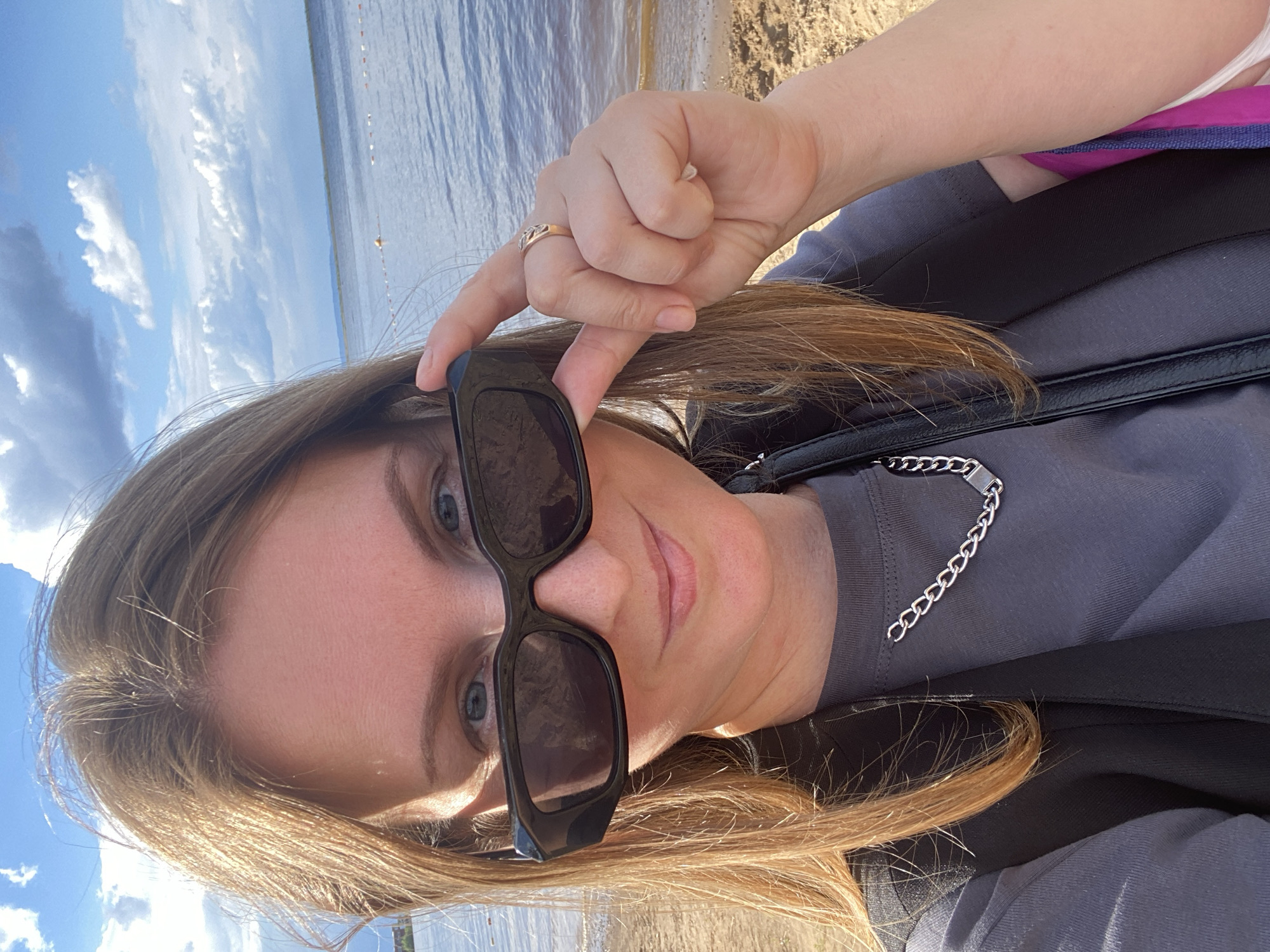 The media file is a close-up, selfie-style portrait of a young woman with long, blonde hair and blue eyes. She is wearing large, black sunglasses and a grey t-shirt with a silver chain detail. A ring is visible on her right hand, which she is using to hold her sunglasses.
The background suggests an outdoor setting, likely near a body of water, possibly a river or lake, with a sandy or dirt shoreline. The sky is a bright blue with scattered white clouds, indicating it is daytime with pleasant weather. The image has a vertical orientation, common for phone-taken selfies.
The woman's expression is neutral, and the overall mood is relaxed and casual, typical of a personal snapshot taken on a day out. There is no visible text in the image. The location context of Chaikovsky, Russia, could imply a riverside park or beach area within or near the city.