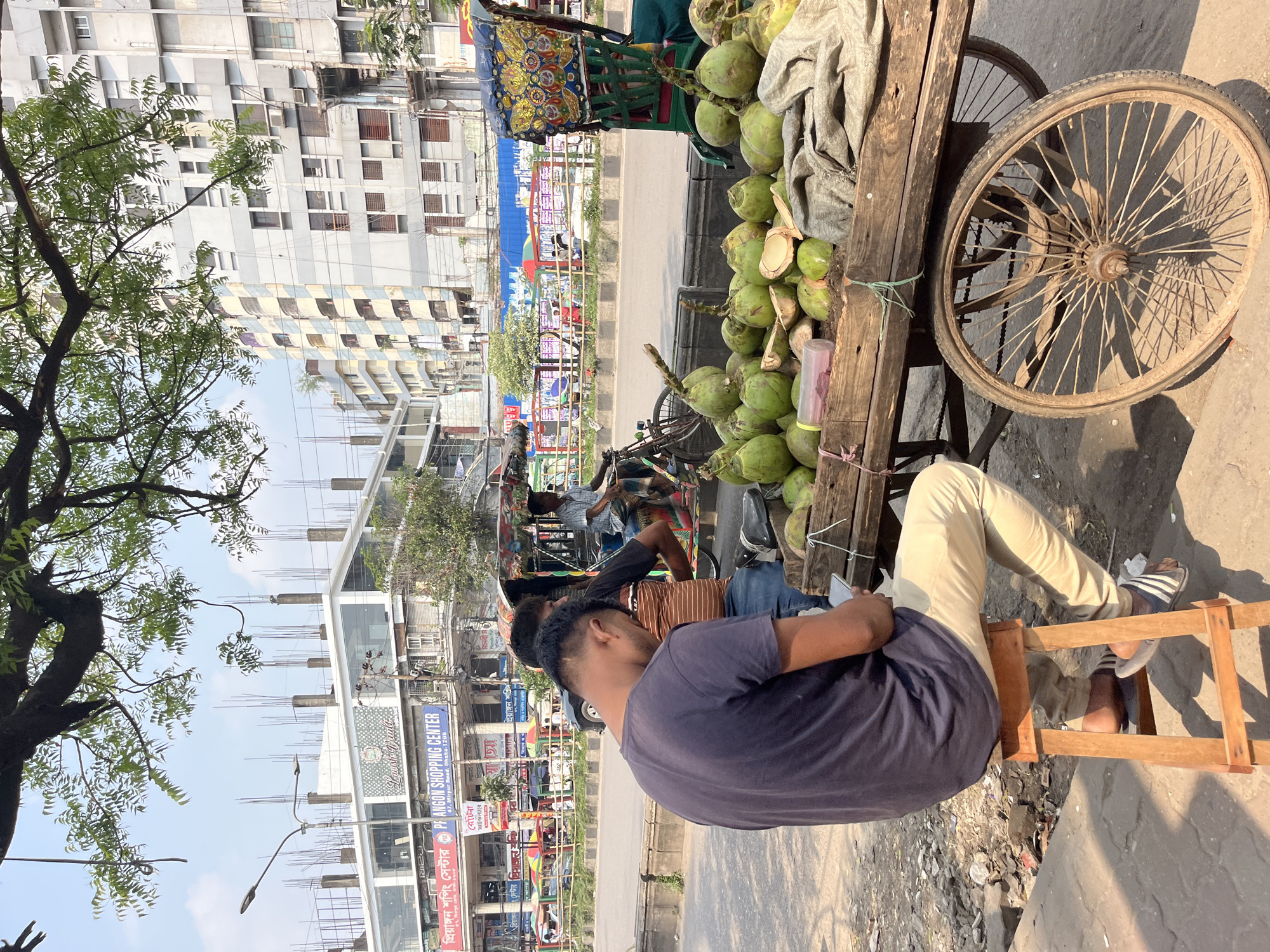 The image depicts a scene in Dhaka, Bangladesh. A man wearing a blue t-shirt and light-colored pants is seated on a simple wooden stool next to a wooden cart laden with green coconuts, some of which are already cut open. The cart has a colorful canopy and a large spoked wheel. The man appears to be looking at or interacting with something in his hands. Behind him are other people, including a man on a rickshaw and other pedestrians. The street is lined with buildings, one of which has signs for "PH ANGON SHOPPING CENTER" and "প্রিয়াঙ্গন শপিং সেন্টার". A large tree with dense foliage is visible on the left side of the image. The setting appears to be a busy street scene.