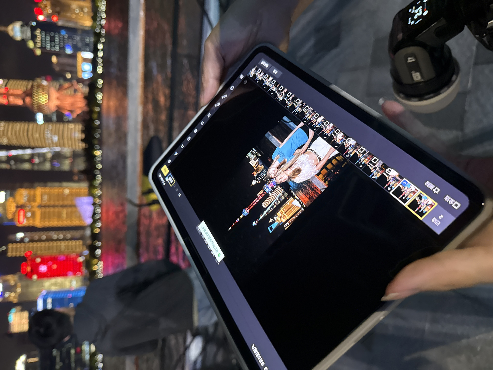 The image captures a close-up view of a tablet device displaying a photograph of two young women posing in front of a brightly lit cityscape at night. The tablet is held by a person whose hand and thumb are visible in the foreground, with a camera-like object attached to its side. The backdrop on the tablet shows iconic skyscrapers and illuminated structures, characteristic of a major metropolitan area like Shanghai. The original image, when viewed in full, reveals a man in a dark hoodie standing in the background, facing away from the camera, with the cityscape also visible behind him. The reflection of the city lights can be seen in the body of water in front of the buildings. The tablet screen shows a video editing interface, with a timeline of images at the bottom. The timestamp on a pop-up window on the tablet reads "9月6日 星期六". While the image does not explicitly state the location as Hongkou District, China, the distinctive skyline with the Oriental Pearl Tower and Shanghai World Financial Center suggests it is the Lujiazui area in Pudong, Shanghai, a very well-known landmark of the city.