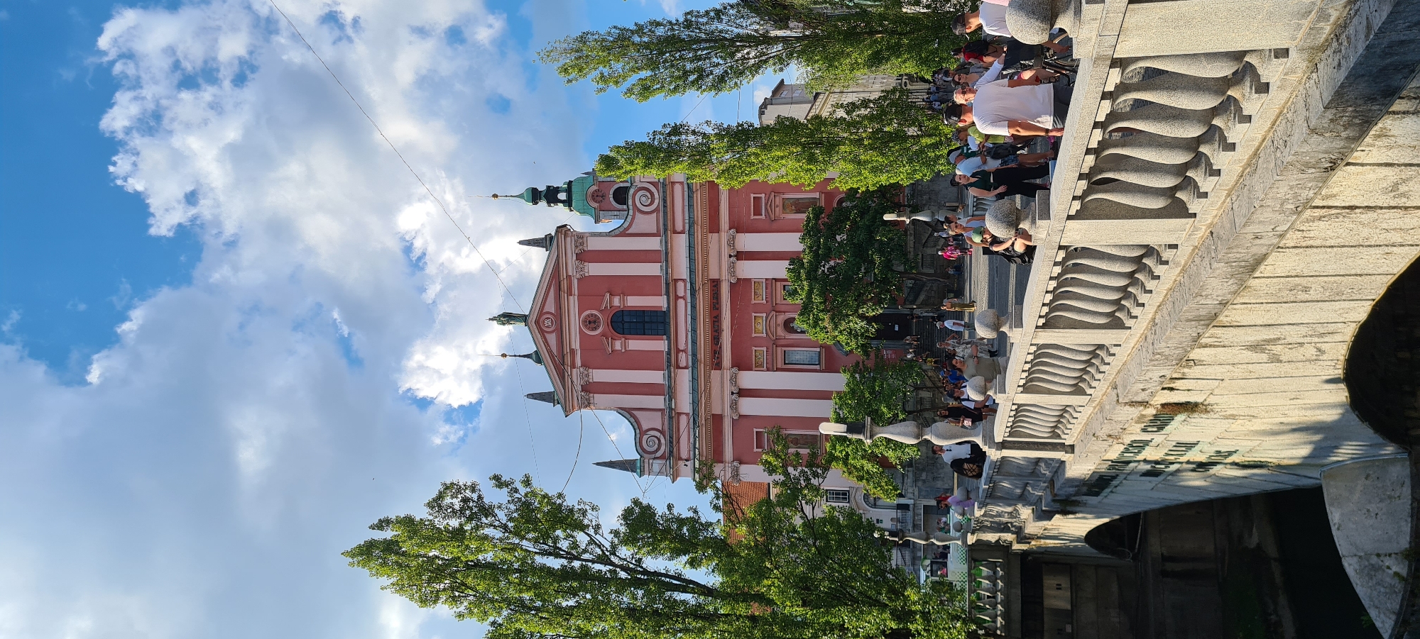 The image captures a daytime view of the Triple Bridge (Tromostovje) in Ljubljana, Slovenia, with the pink facade of the Church of St. Francis of Assisi in the background. The bridge, made of stone and adorned with ornate balustrades, spans a waterway. A crowd of people is visible on the bridge and in the square in front of the church. Tall, slender trees line the sides of the square. The sky is blue with scattered white clouds. The scene is bathed in sunlight, casting shadows and highlighting the architectural details of the bridge and the church.