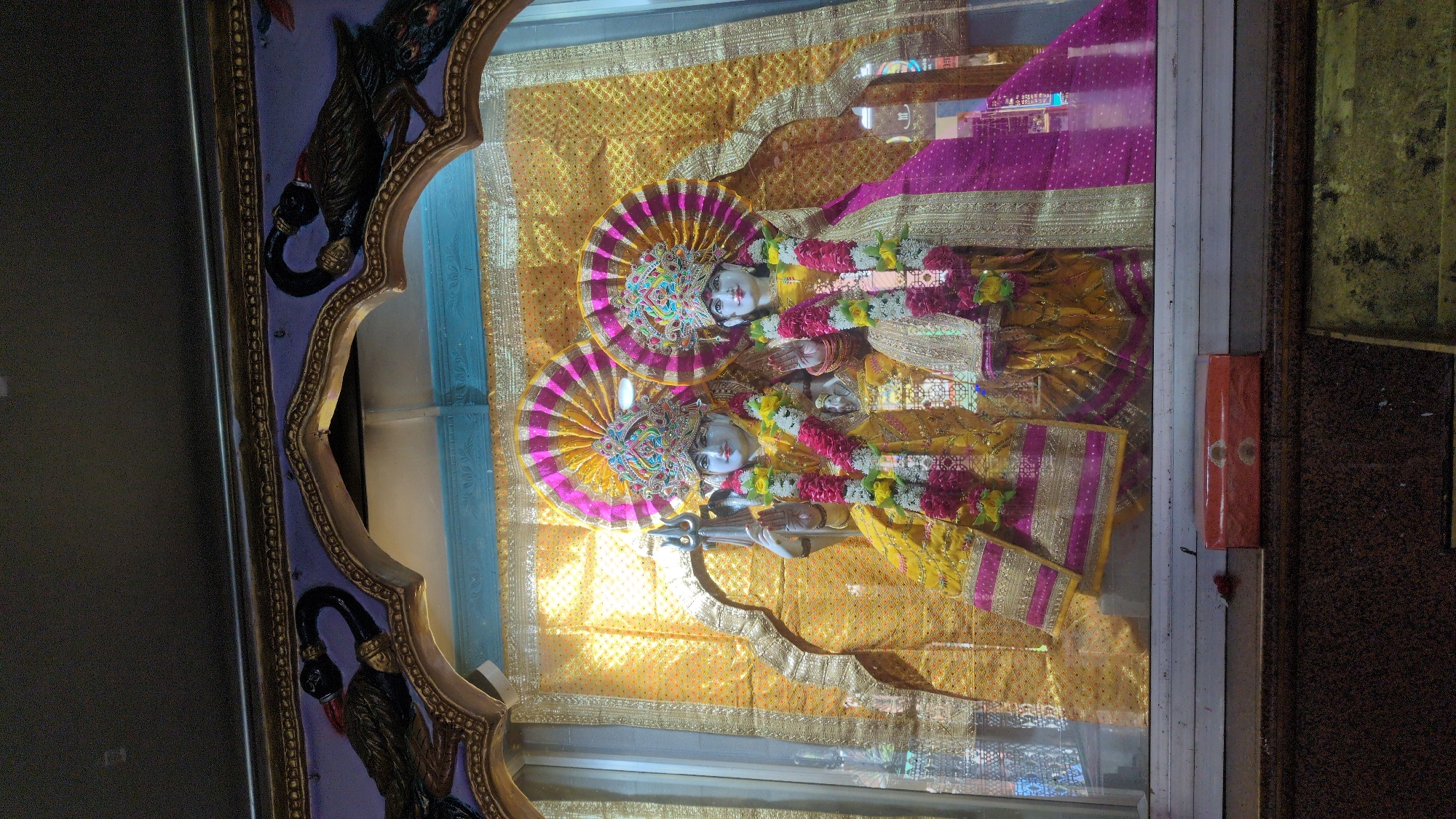 This image captures a devotional scene featuring two elaborately adorned deities, likely idols or statues, prominently displayed within what appears to be a Hindu temple or shrine in Bhagsu, India. The deities are positioned centrally, facing the viewer, and are richly decorated.
Both figures wear vibrant yellow and pink/magenta garments with ornate gold borders and intricate patterns. Their heads are crowned with magnificent, multi-colored headdresses featuring beadwork and decorative elements. Multiple garlands of fresh flowers, in shades of white, yellow, and pink/red, drape across their chests and shoulders. The deity on the left (as viewed when correctly oriented) holds a silver trident (trishula) in their lower right hand, while their upper hands are held in a gesture of blessing or welcome. The deity on the right displays a similar hand gesture.
They stand before a backdrop of shimmering golden fabric adorned with intricate patterns, creating an opulent setting. The entire display is enclosed behind a clear glass panel, which reflects ambient light and portions of the surroundings. To the left of the main display, an ornate archway with a purple-blue background and gold trim features dark, sculpted figures, possibly mythological creatures or elephants, adding to the decorative grandeur. To the right, a simple white frame borders the display, beyond which lies a darker, textured wall and an orange rectangular object.
The scene is brightly illuminated, creating a reverent and spiritual atmosphere. The detailed craftsmanship of the idols, their attire, and the surrounding decorations are characteristic of Hindu devotional art. No discernible text is visible in the image.