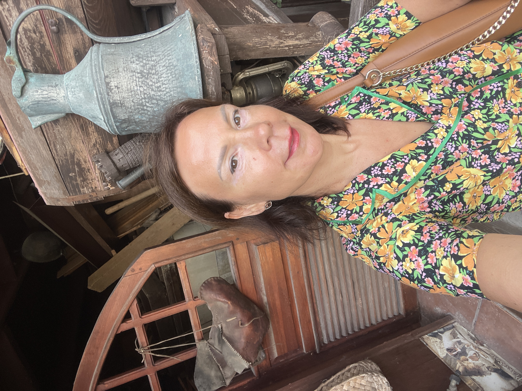 A woman with shoulder-length dark hair and makeup is taking a selfie. She is wearing a black, floral-print shirt with green trim and a brown leather purse with a gold chain strap over her right shoulder. She is standing in front of a wooden structure with various antique items, including a large, aged pitcher, a lantern, and a wooden shoe. The background suggests an outdoor setting, possibly a market or antique shop, with a wooden window frame and a woven basket visible. The lighting is bright, indicating daytime. The location is identified as Sochi, Russia.