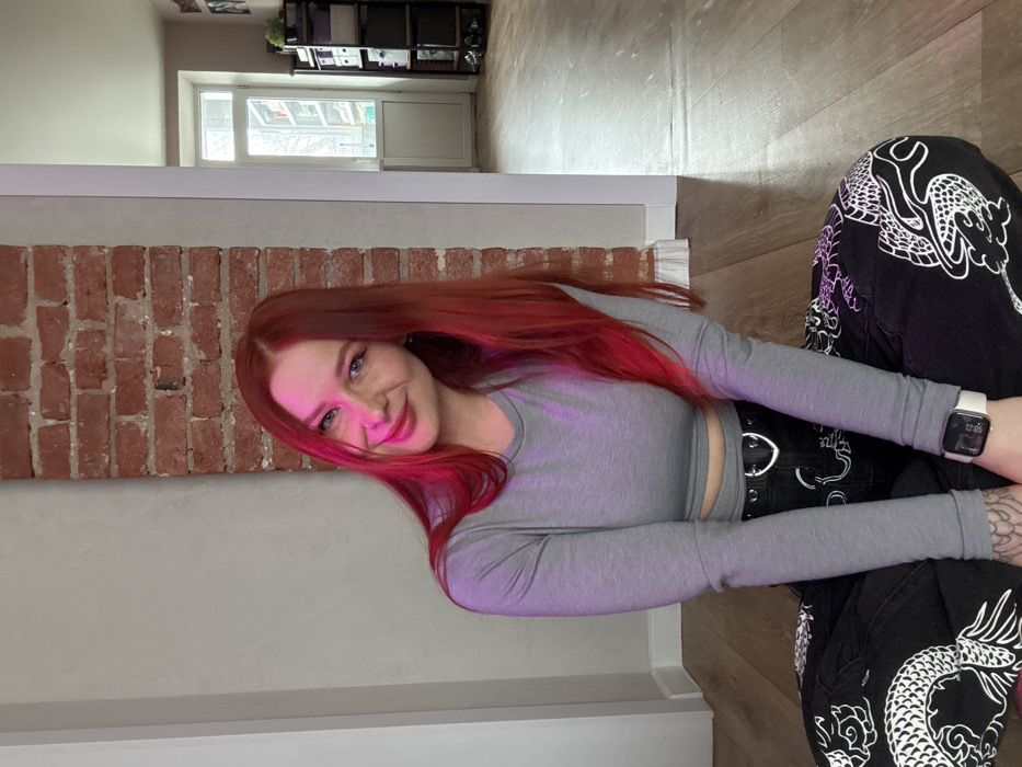 The image depicts a female individual seated cross-legged on a light brown wooden floor in an interior space in Rostov-on-Don, Russia. The subject, a young adult with long, auburn hair, wears a grey long-sleeved crop top, black pants adorned with white ornate patterns, a black belt featuring a silver D-ring buckle, and a white smartwatch on her left wrist. Tattoos are visible on her right hand. She is positioned facing forward, smiling towards the camera, with the left side of her face and hair illuminated by a distinct pink-purple light source. The background consists of a light grey wall to the subject's left, a vertical section of exposed red brickwork behind her, and another light grey wall to her right. Further in the background, a white-framed doorway is visible on the right, revealing a window with indistinct outdoor elements and a dark shelving unit holding various objects.
