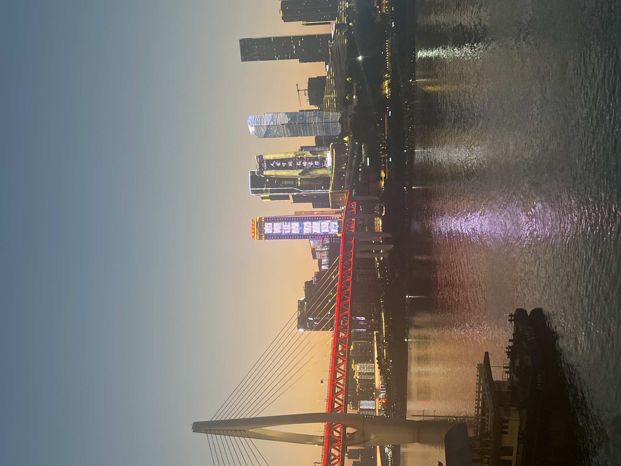 This is an image of the Chongqing skyline at dusk, focusing on the Jialing River. In the foreground, a portion of a dark-colored boat is visible on the left, its hull low in the water. The river occupies the lower half of the image, reflecting the city lights with hues of orange, yellow, and purple.
The middle ground features a modern suspension bridge with a tall white pylon on the left. The bridge's deck is illuminated with vibrant red lights, creating a strong horizontal line across the scene. Behind the bridge, the city's skyline is dominated by several tall skyscrapers. Some of these buildings are lit with colorful displays, including vertical lines of white and purple lights on the far left building and a prominent display of Chinese characters on a central building. Other buildings are illuminated with white and yellow lights. Construction cranes are visible between some of the skyscrapers.
The background consists of a clear sky transitioning from a light blue at the top to a warm, soft orange near the horizon, indicating the setting sun. The overall atmosphere is urban and modern, with a notable contrast between the natural elements of the river and sky and the constructed urban environment.