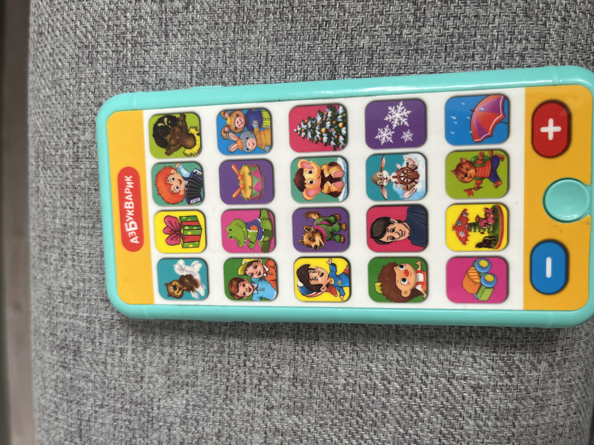 The image shows a close-up of a children's toy phone, likely an alphabet learning device, placed on a textured gray surface, possibly a couch or fabric mat. The toy phone is predominantly turquoise and yellow. It features a grid of colorful square buttons, each depicting a cartoon character or object. The top of the phone has a yellow banner with Russian text that reads "АЗБУВАРИК," which translates to "ABC book" or "Alphabet book." The grid contains various images including animals like a bear, a squirrel, a crocodile, a dog, a monkey, rabbits, and a cat, as well as children, a gift box, a drum, a Christmas tree, snowflakes, a car, a carousel, and an umbrella in the rain. At the bottom of the phone are three larger buttons: a round turquoise button with a white dash, a circular turquoise button with a white center, and a red oval button with a white plus sign. The background is a soft focus gray texture. The toy appears to be new and in good condition. The location in Poronaysk, Russia is not discernible from the image.