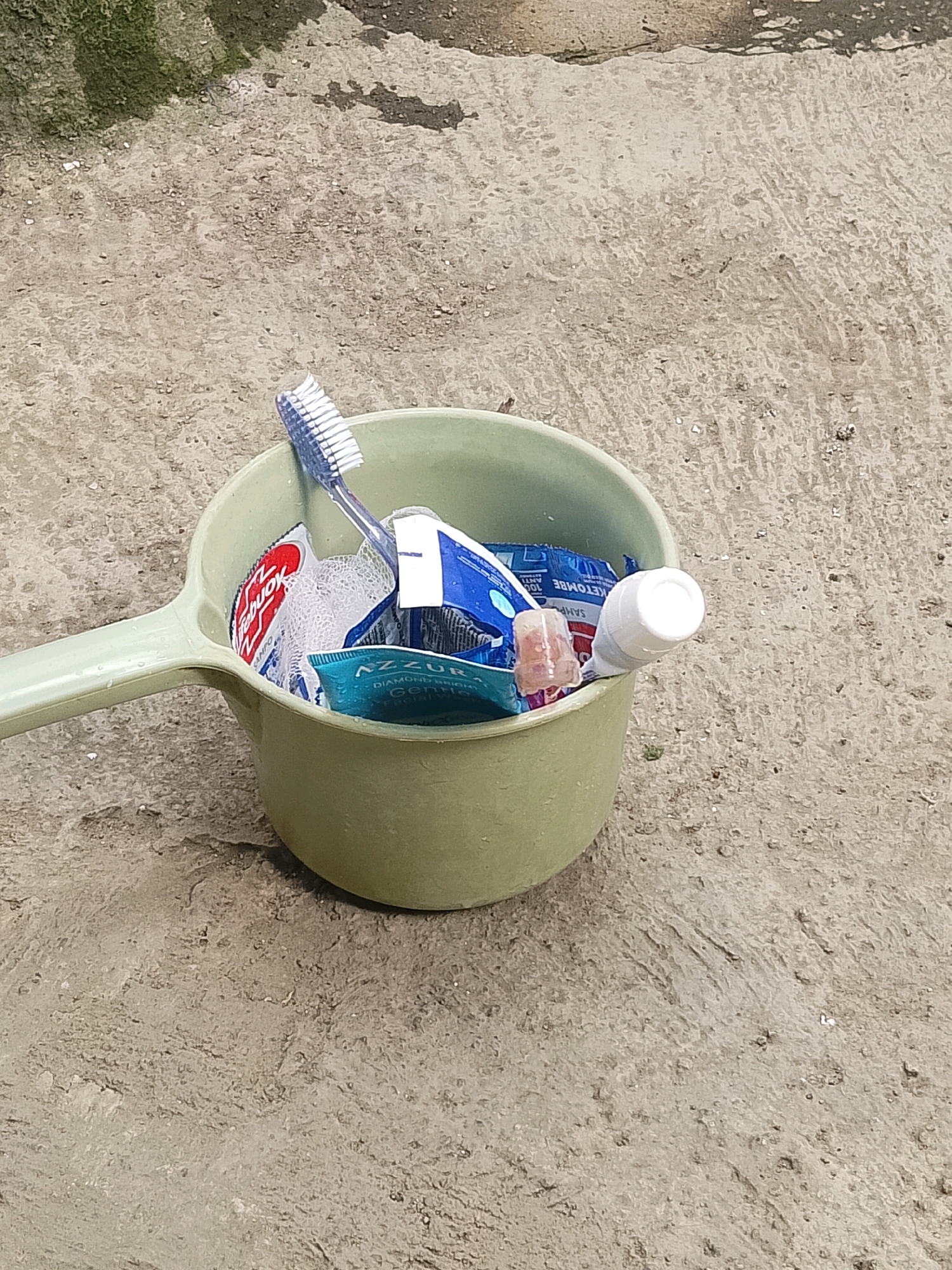A light green plastic dipper filled with toiletries sits on a rough concrete surface. The dipper contains a blue and white toothbrush, a white mesh soap scrubber, a tube of Colgate toothpaste, a small tube of facial wash labeled "AZZURRA DIAMOND BRIGHT Gentle Facial Wash," and a small white tube with a cap. The concrete surface appears weathered and is speckled with small dark particles. In the upper left corner of the image, a patch of damp, dark green moss is visible on another section of concrete. The scene appears to be outdoors, possibly in an alley or a less-maintained area. The lighting suggests it is daytime.